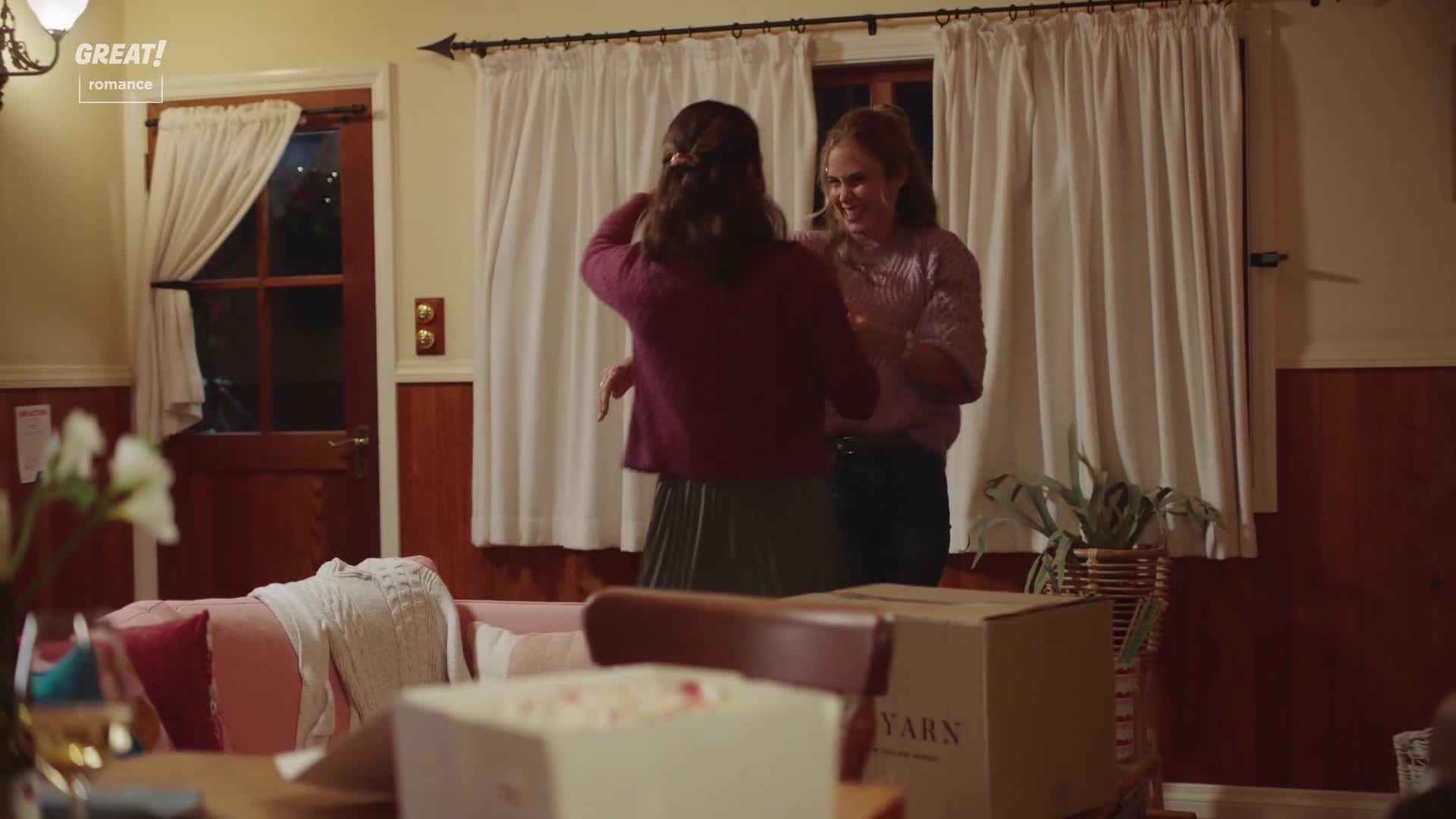 Two women are laughing, arms raised as if they're embracing. The room is cozy, with a sofa and a box labeled "Yarn" nearby.
