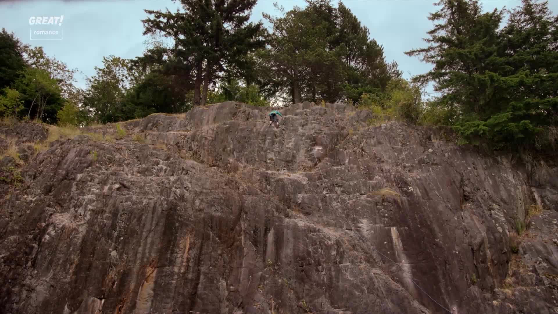 A figure, small against the rock face, is slowly making their way up. The grey stone is textured with lines, and the trees at the top offer a cool contrast to the climb.
