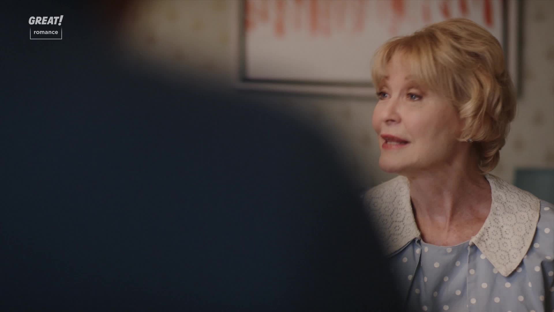 A woman with blonde hair and a patterned dress is speaking, her expression animated. The scene feels like a cozy, domestic setting, perhaps a moment from a British romance.
