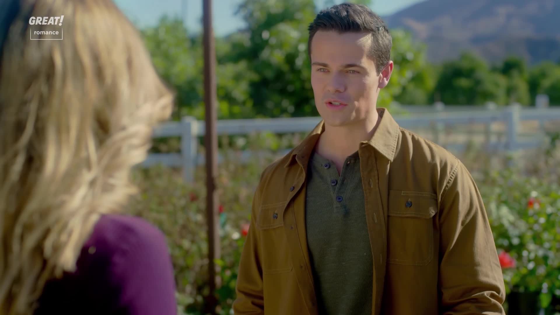 He's speaking, looking at a woman with blonde hair, his expression earnest. The setting is a sunny, outdoor space with a white fence and flowers in the background.
