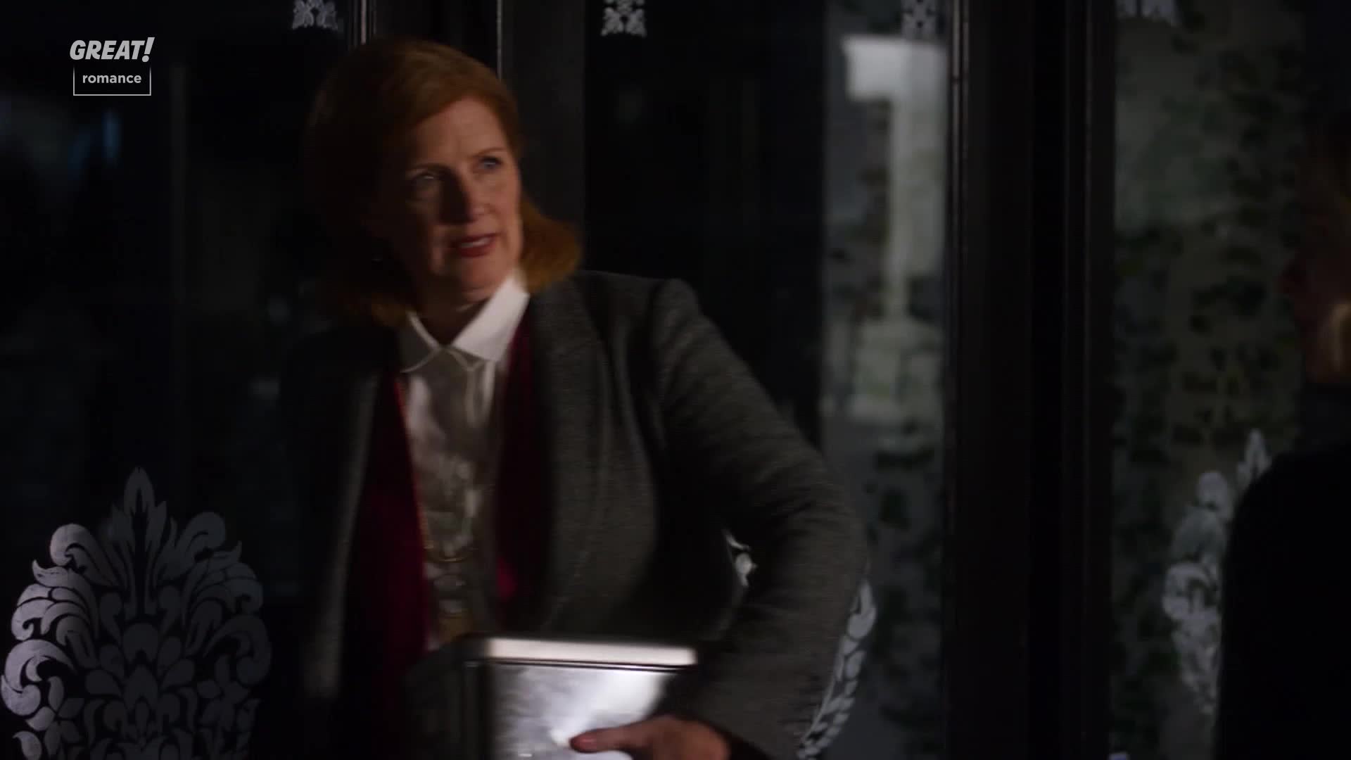 A woman in a grey blazer emerges from a doorway, holding a metal container. She glances to her right, a slight smile playing on her lips.
