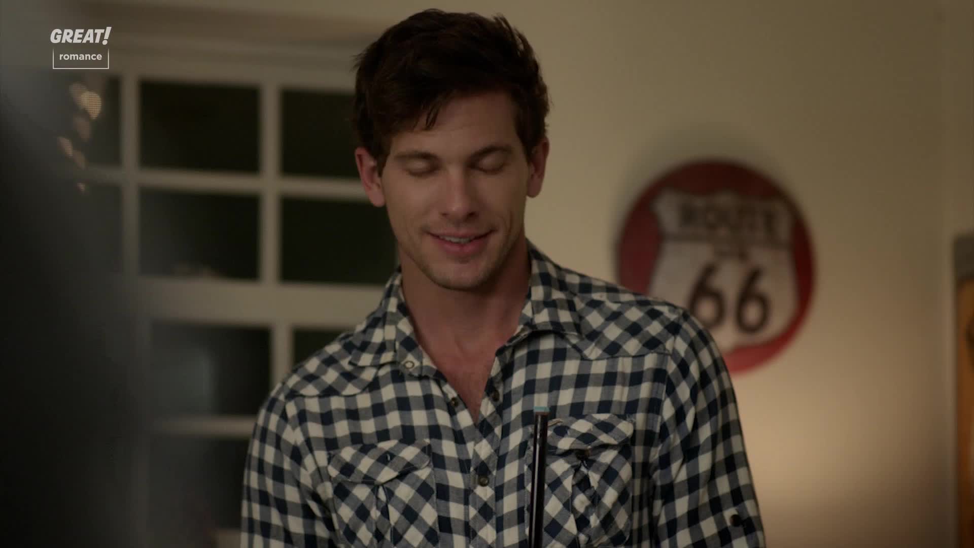 The man in the checkered shirt smiles, his eyes crinkling slightly. Behind him, a Route 66 sign hangs on the wall.
