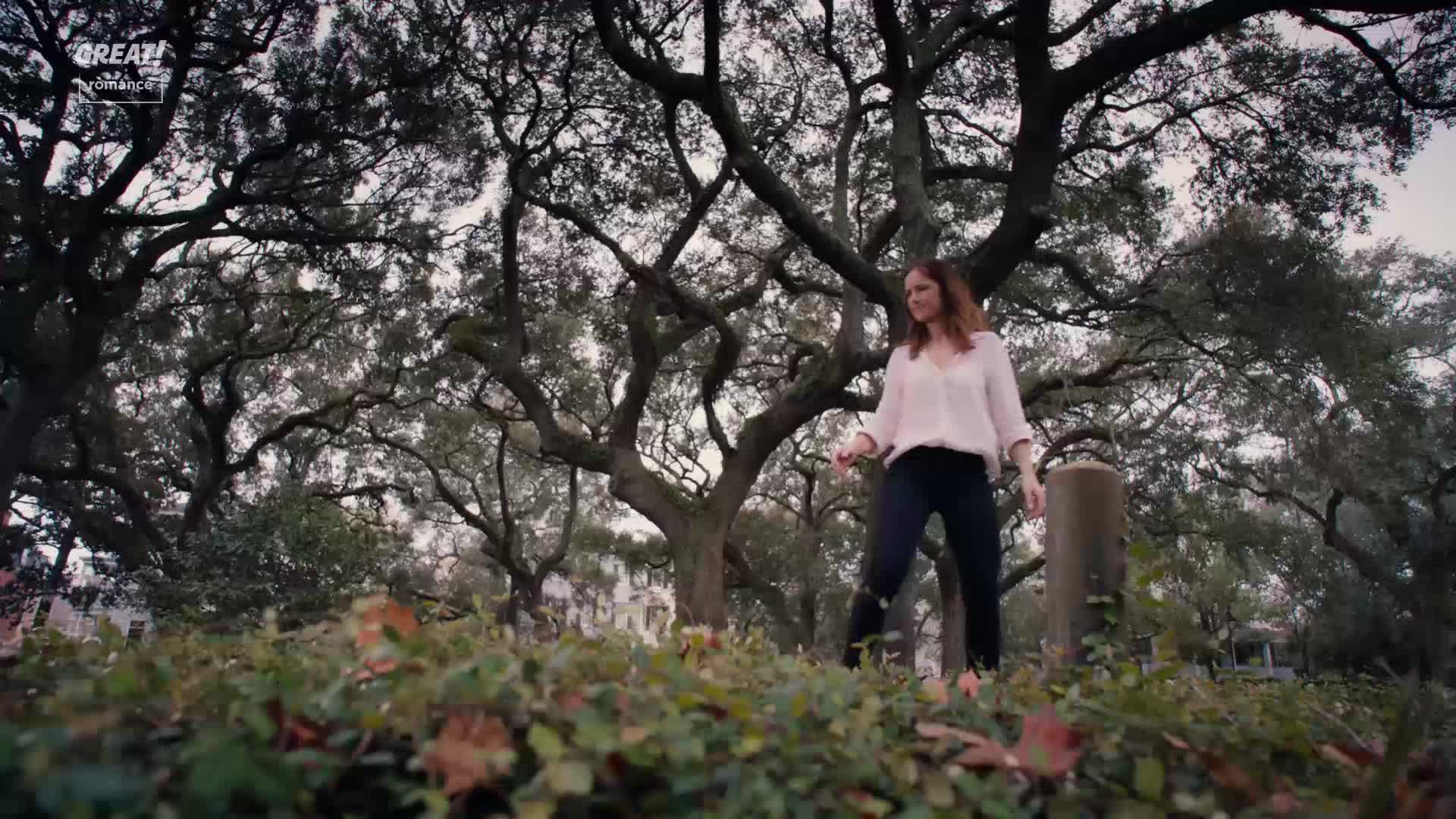 A woman in a light pink blouse and dark trousers stands in a park, looking down. The branches of a large tree spread overhead, framing the sky.
