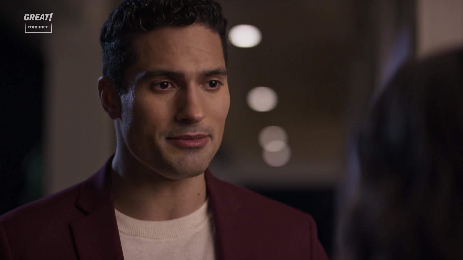 He's looking at someone, a slight smile playing on his lips. The maroon jacket looks sharp against his pale shirt.
