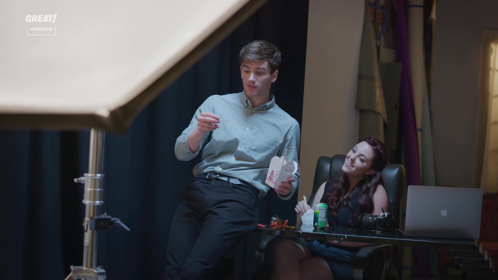 A man, holding a takeaway container, gestures as he speaks. Across from him, a woman smiles, leaning back in her chair at a desk cluttered with a laptop and camera.
