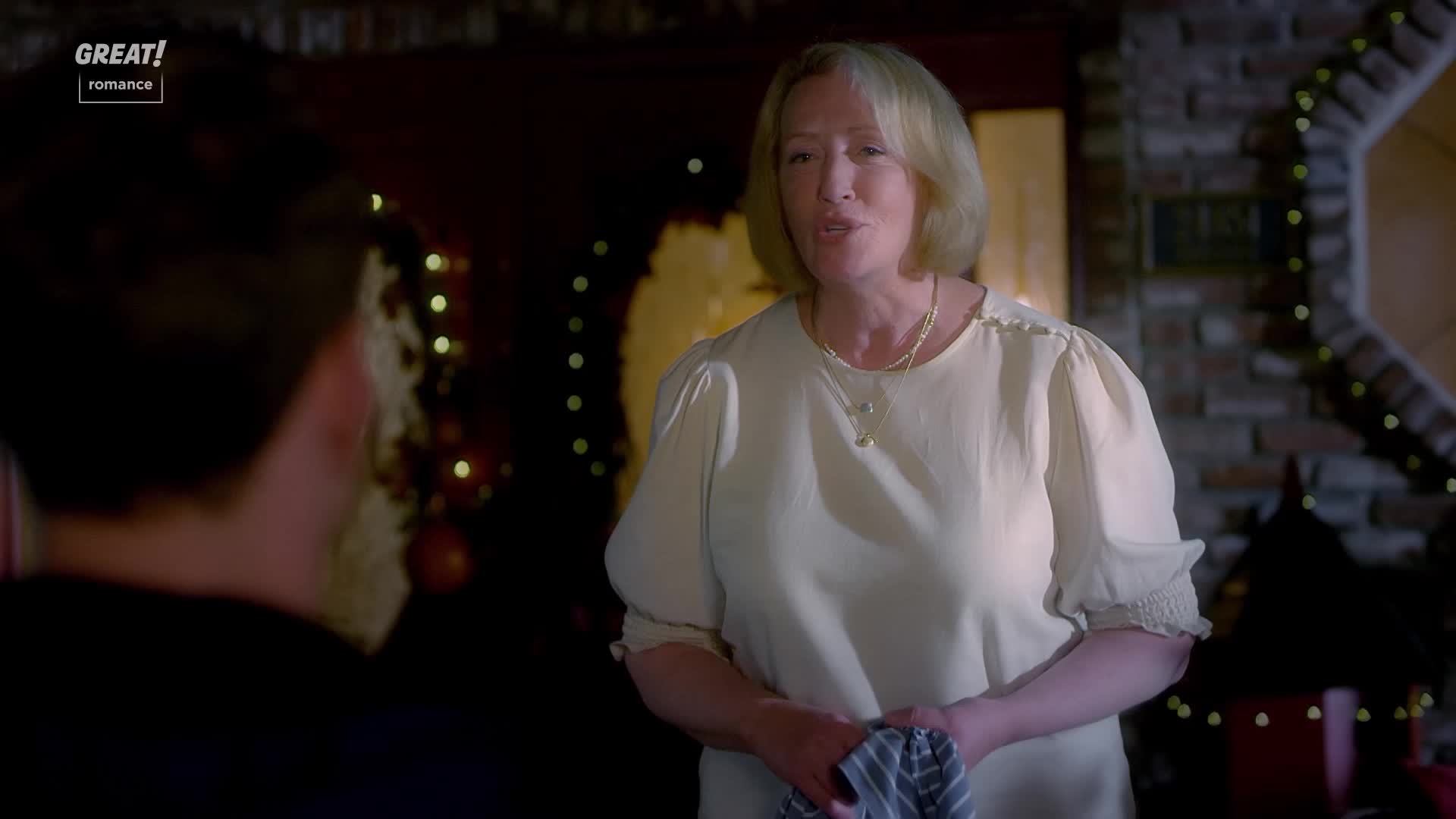 A woman with blonde hair and a cream-colored blouse is talking, a tea towel clutched in her hands. Behind her, a brick fireplace is adorned with twinkling lights.

