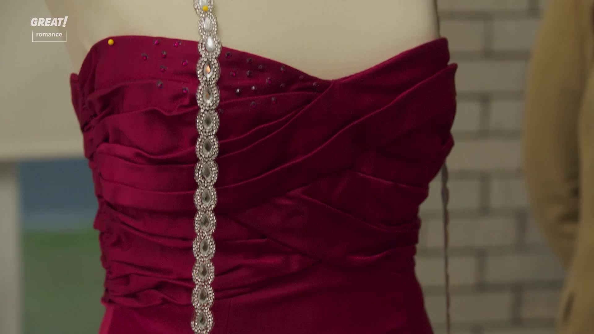 A deep red, strapless dress is draped on a mannequin, with a sparkling, jeweled trim hanging down the front. The fabric is gathered and pinned, suggesting a bespoke design in progress.
