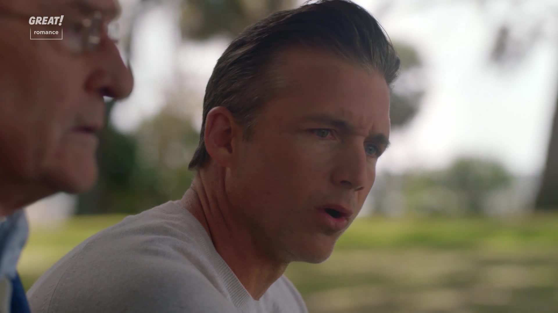 A man with slicked-back hair stares intently, his mouth slightly open as if he's about to speak. Beside him, an older man in glasses listens, his expression thoughtful.
