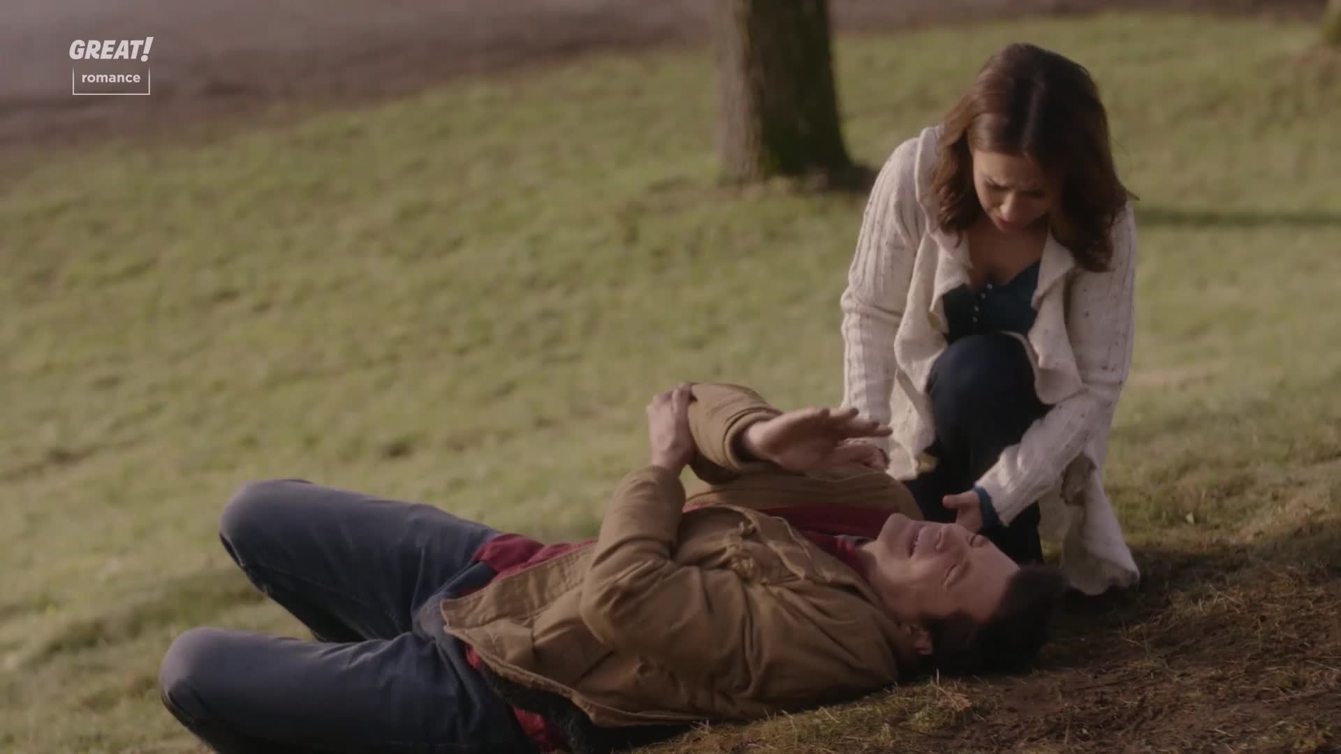 A woman kneels beside a man lying on the grass, her hand hovering over his chest. He's looking up at her, his face etched with concern.
