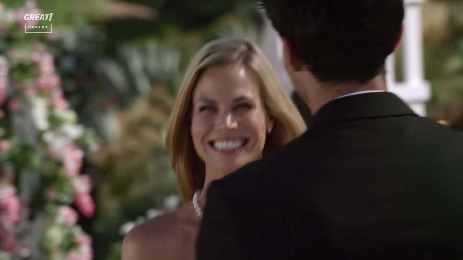 A woman in a white dress beams at the man in front of her, a pearl necklace gleaming at her neck. The man's back is turned, his black suit a stark contrast to the soft, floral background.

