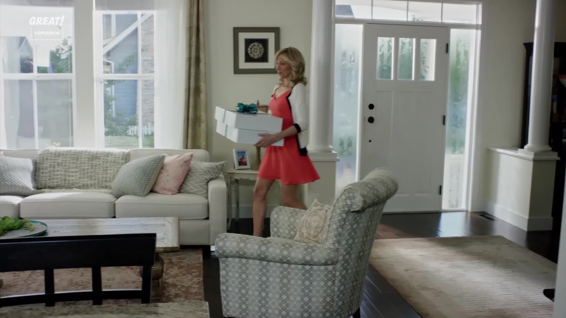 A woman in a bright red dress strides through the living room, carrying a stack of white boxes. She's heading toward the front door, likely on her way out.
