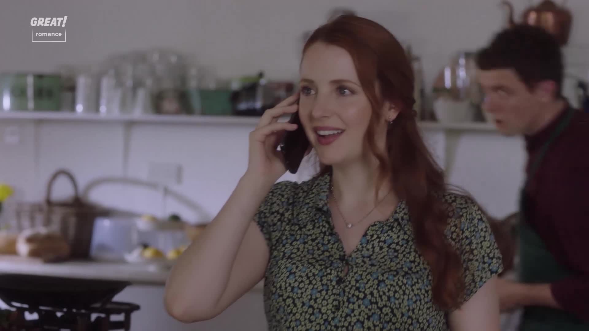 A woman with red hair smiles as she chats on her phone in a sunlit kitchen. Behind her, a man in an apron looks on with a surprised expression.
