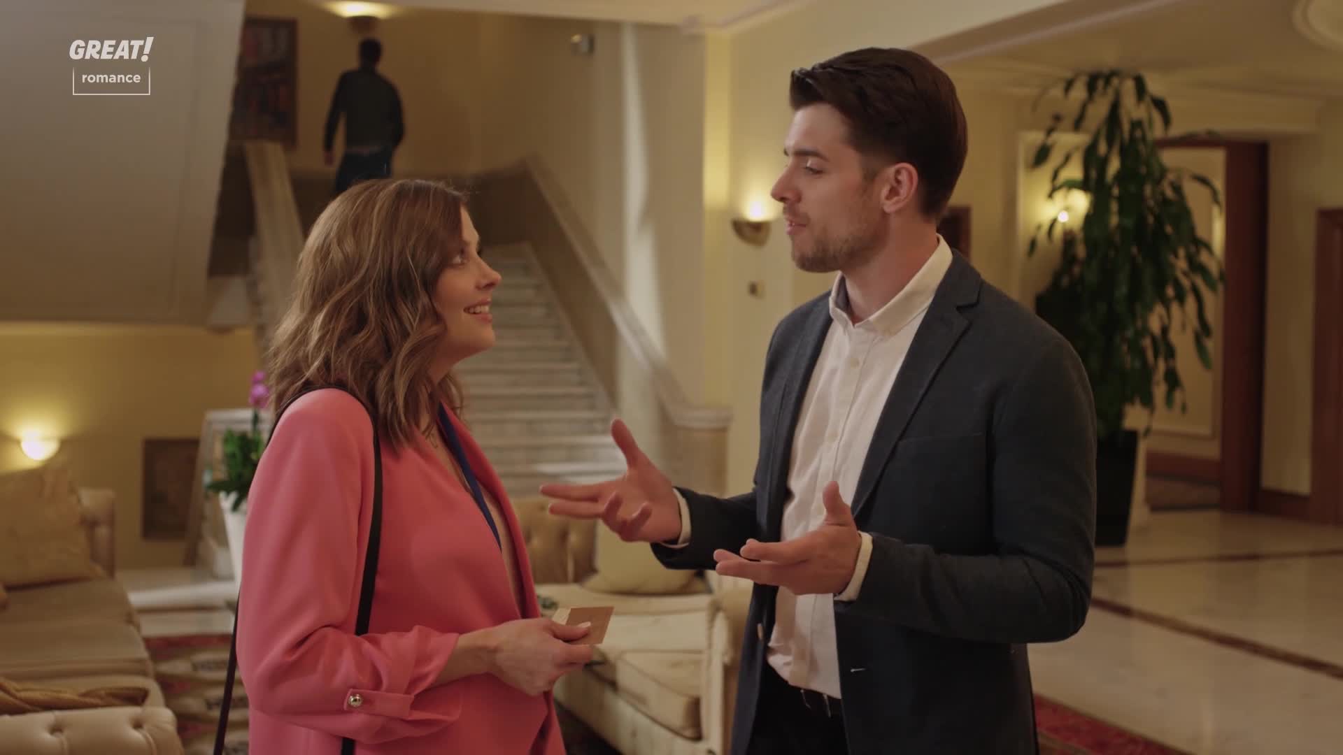 A man in a dark blazer gestures animatedly, speaking to a woman in a pink jacket. She holds a card, smiling, while a figure walks up a grand staircase in the background.
