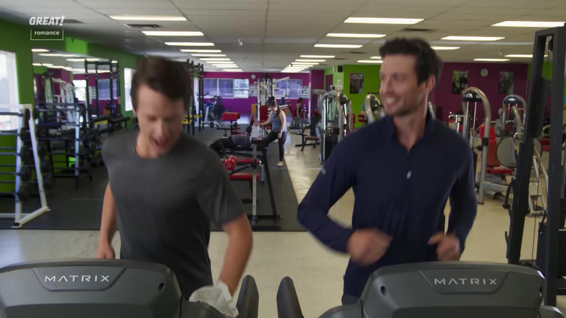 Two men, both smiling, are jogging on treadmills side-by-side in a gym. One of them is holding a white cloth, maybe to wipe his face, as they run.
