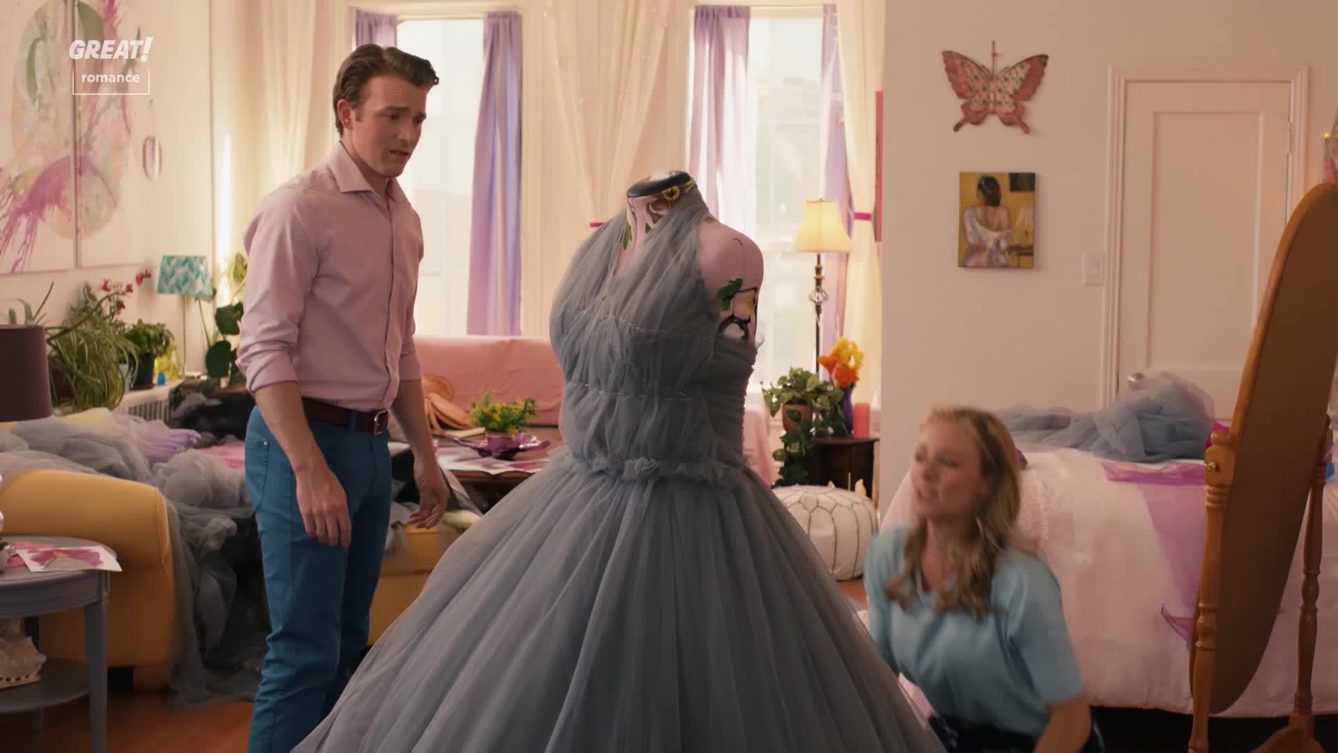A man in a pink shirt and jeans stares at a large, grey gown on a dress form. The woman on the floor looks up, smiling, as if she's admiring the dress.
