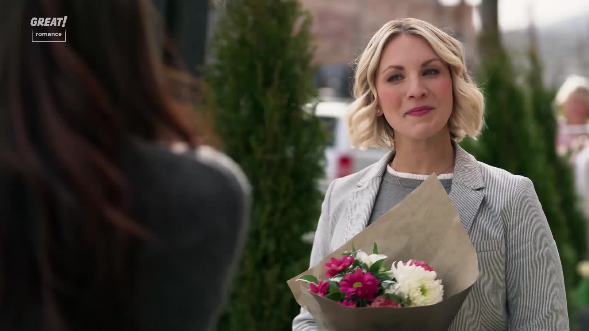 The woman with the blonde bob holds a bouquet, her eyes focused on someone out of frame. She's wearing a grey blazer and a soft smile, the flowers a vibrant splash of color against her outfit.
