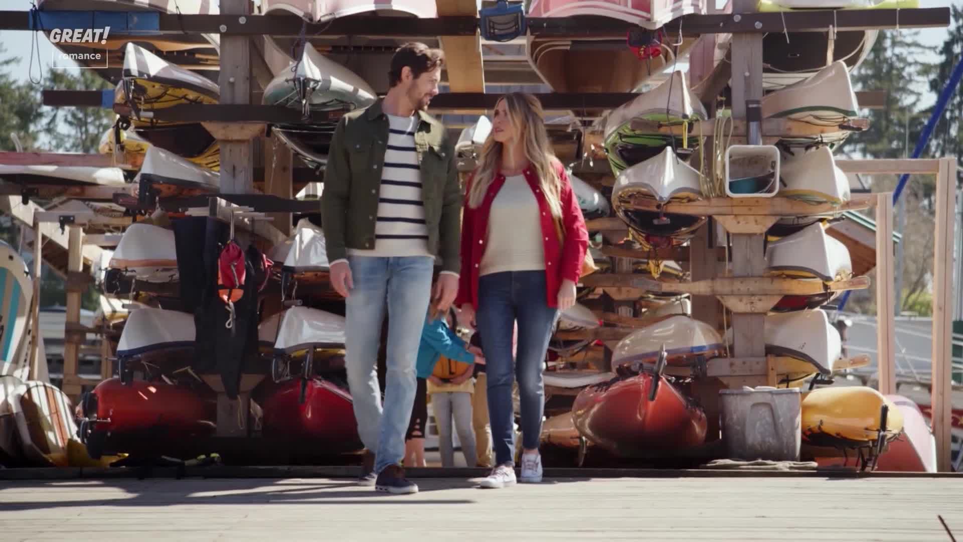A couple strolls along a wooden dock, holding hands as they look at each other. Behind them, a rack of boats in various colors is stacked, suggesting a scenic location in the United Kingdom.
