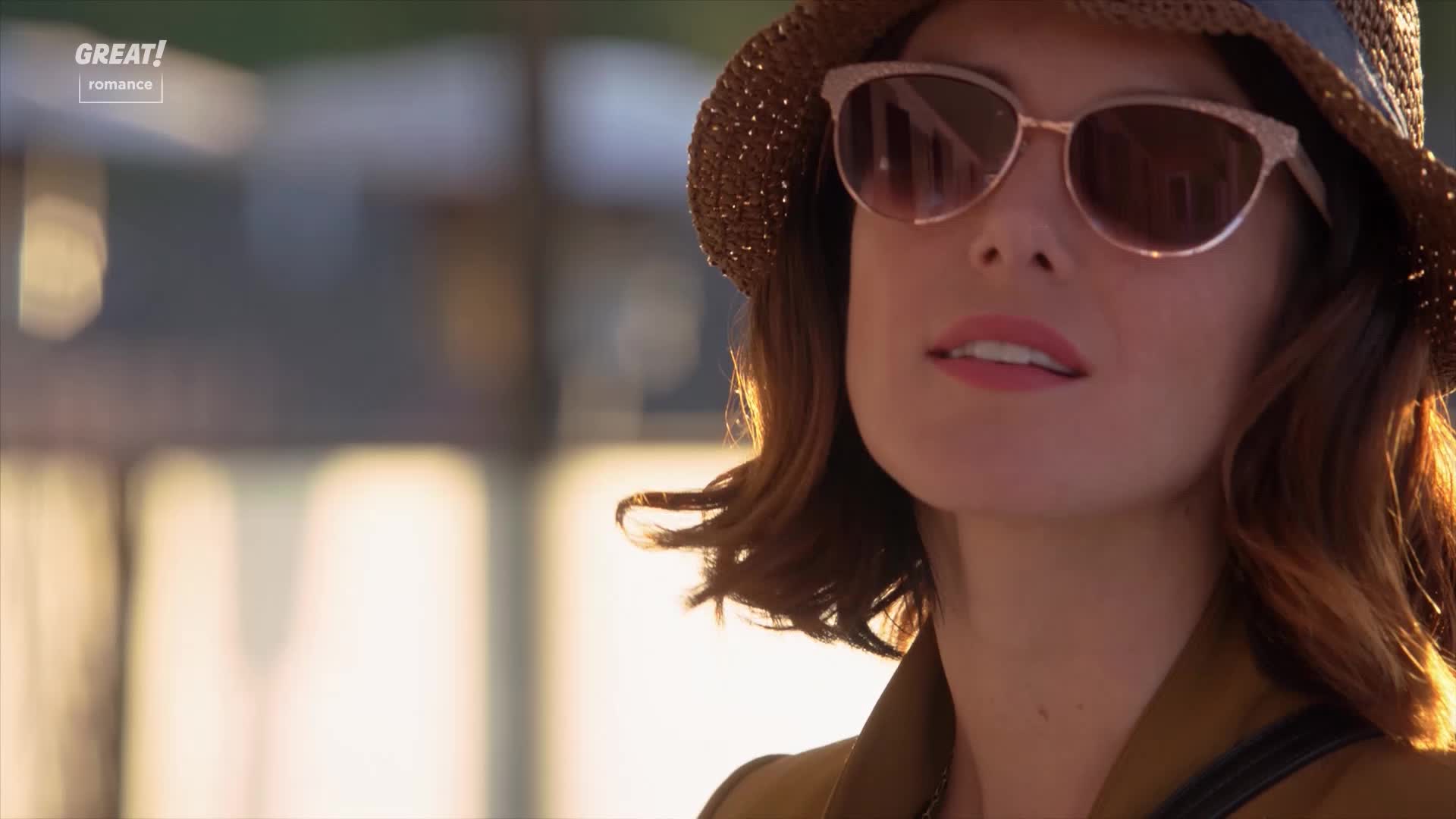 Sunlight catches the woman's hair, highlighting the gentle waves. She's wearing sunglasses and a hat, and her gaze is soft.
