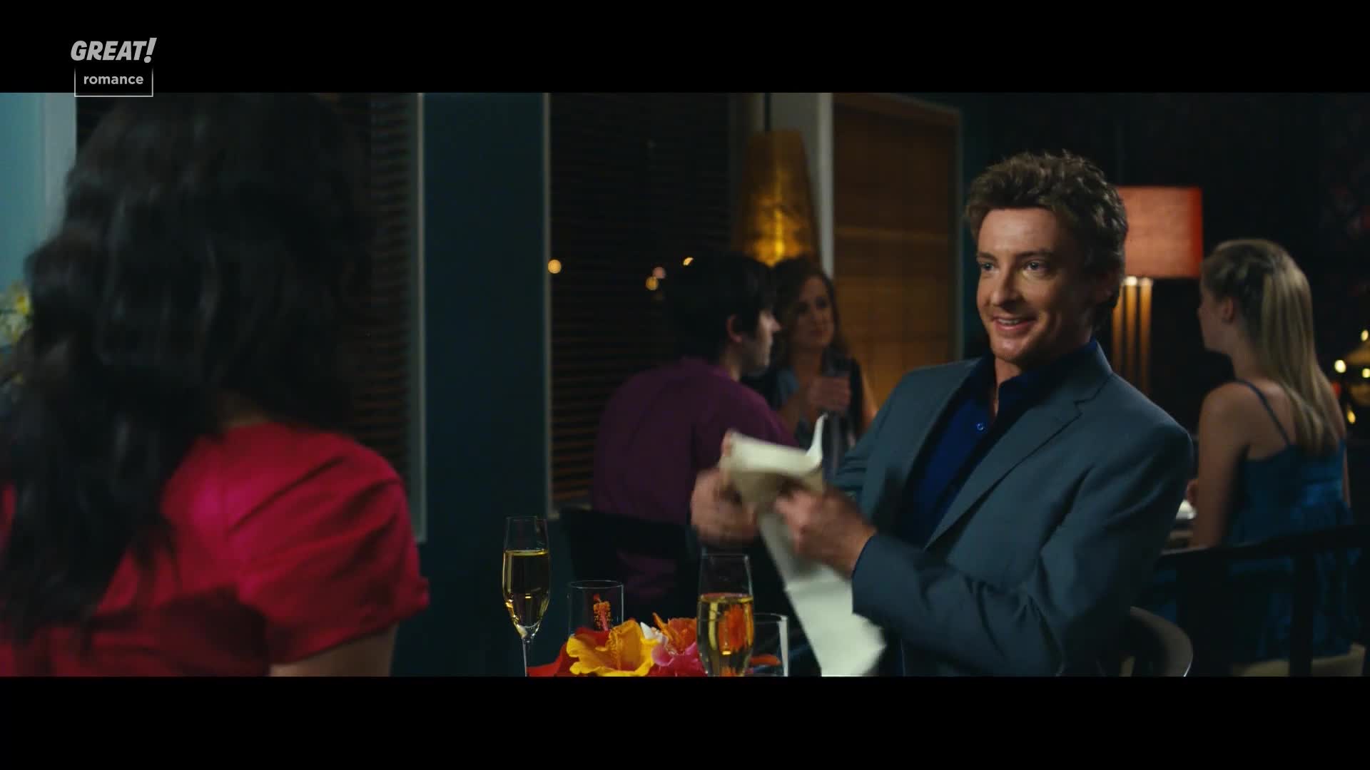 A man in a blue suit holds a napkin, smiling at someone across the table. Other diners chat in the background, the scene lit by warm lamps and soft candlelight.
