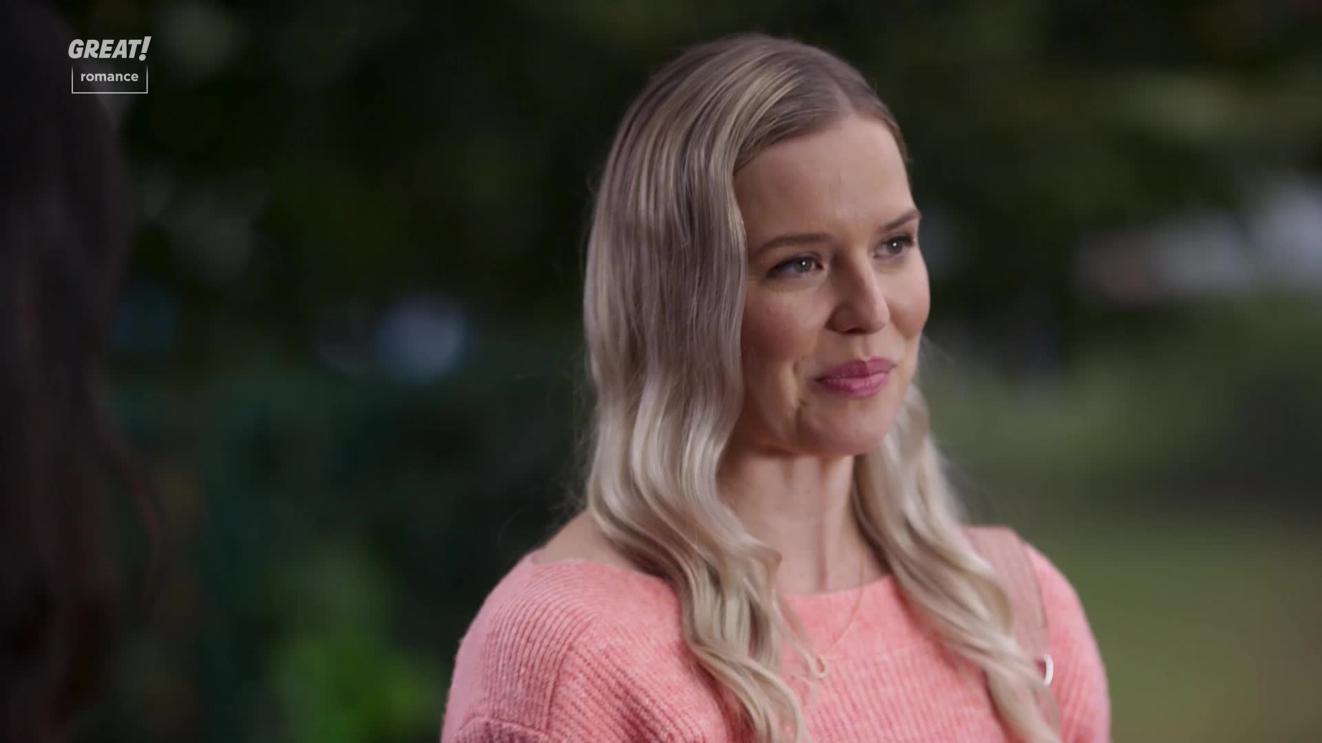 A woman with wavy blonde hair smiles softly, her gaze directed off-screen. She's wearing a pink sweater, and a necklace subtly catches the light.
