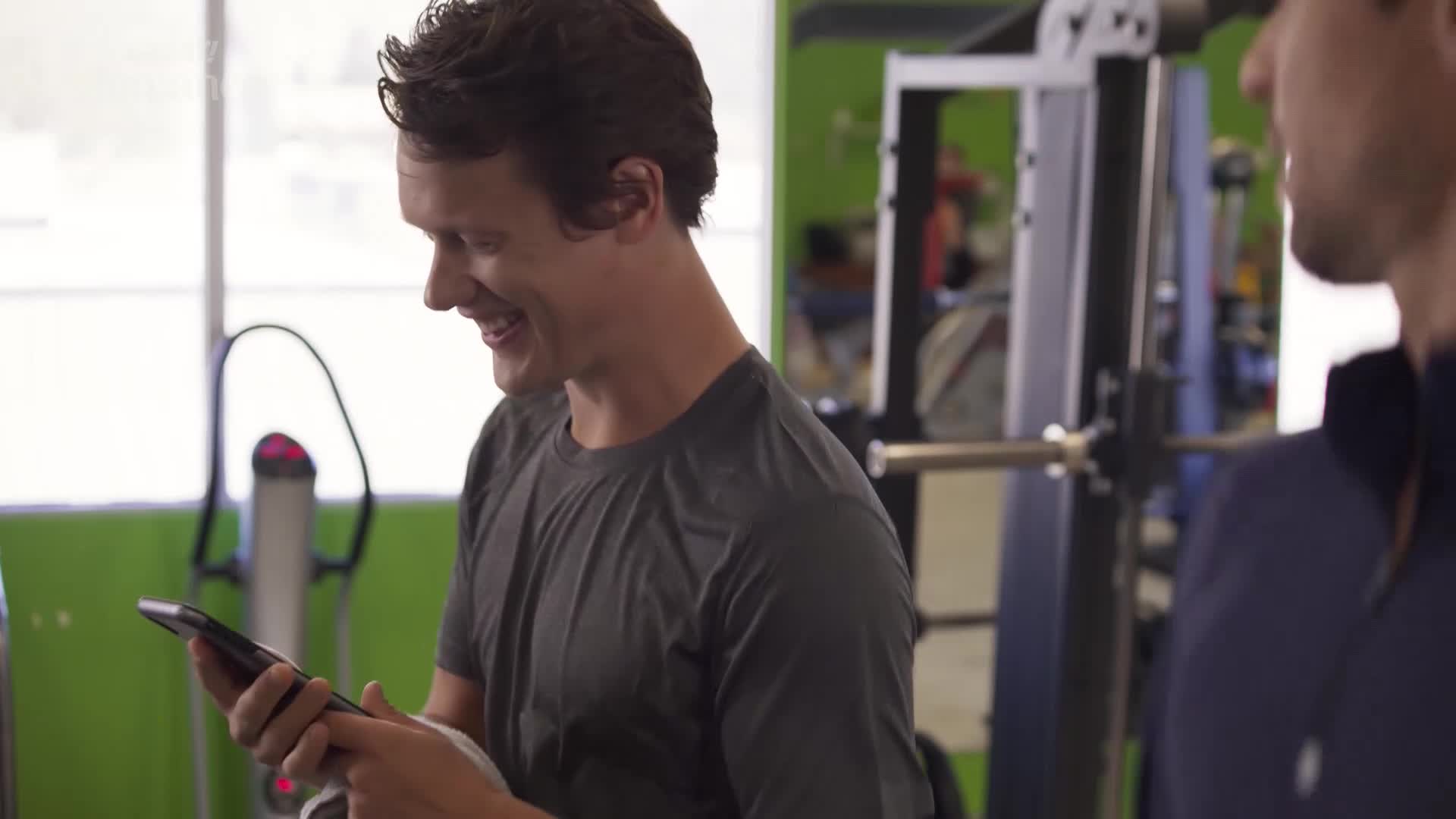 A man in a grey t-shirt is smiling, looking down at his phone. He's standing in a gym, and another man is partially visible beside him.
