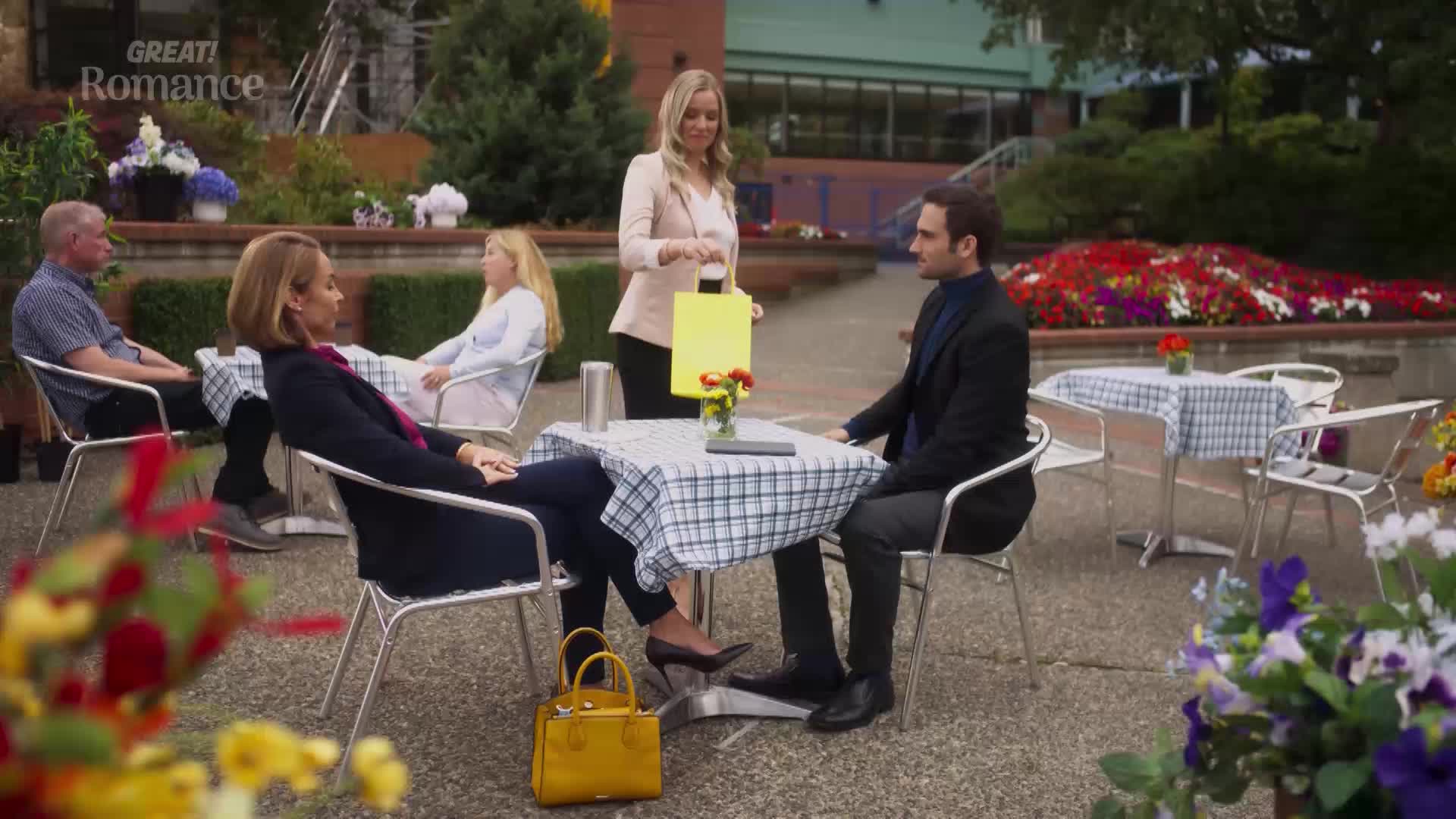 A woman in a blazer presents a yellow notepad to a couple seated at a table, while other patrons relax nearby. The scene unfolds outdoors, surrounded by flowerbeds and a hint of a building in the background.
