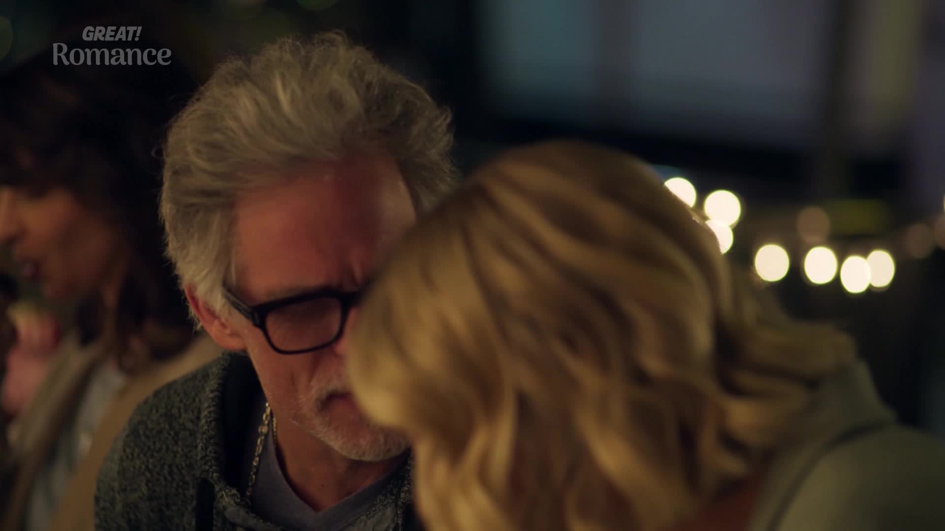 A man in glasses leans close to a woman, their faces nearly touching. Soft light from behind them illuminates the woman's blonde hair, highlighting the intimate moment.
