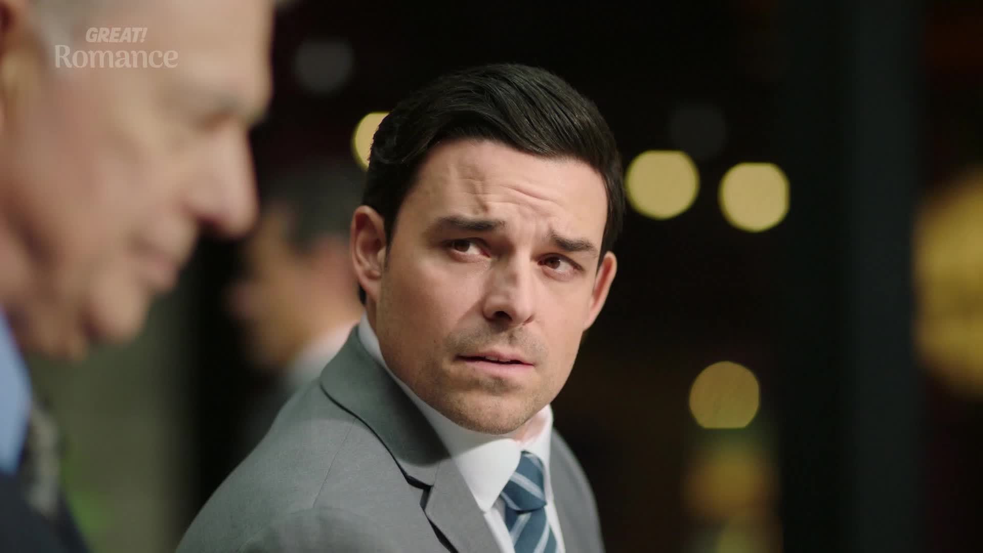 The man in the grey suit looks concerned, his brow furrowed as he listens to someone off-camera. His eyes are wide, and he seems to be processing something important.
