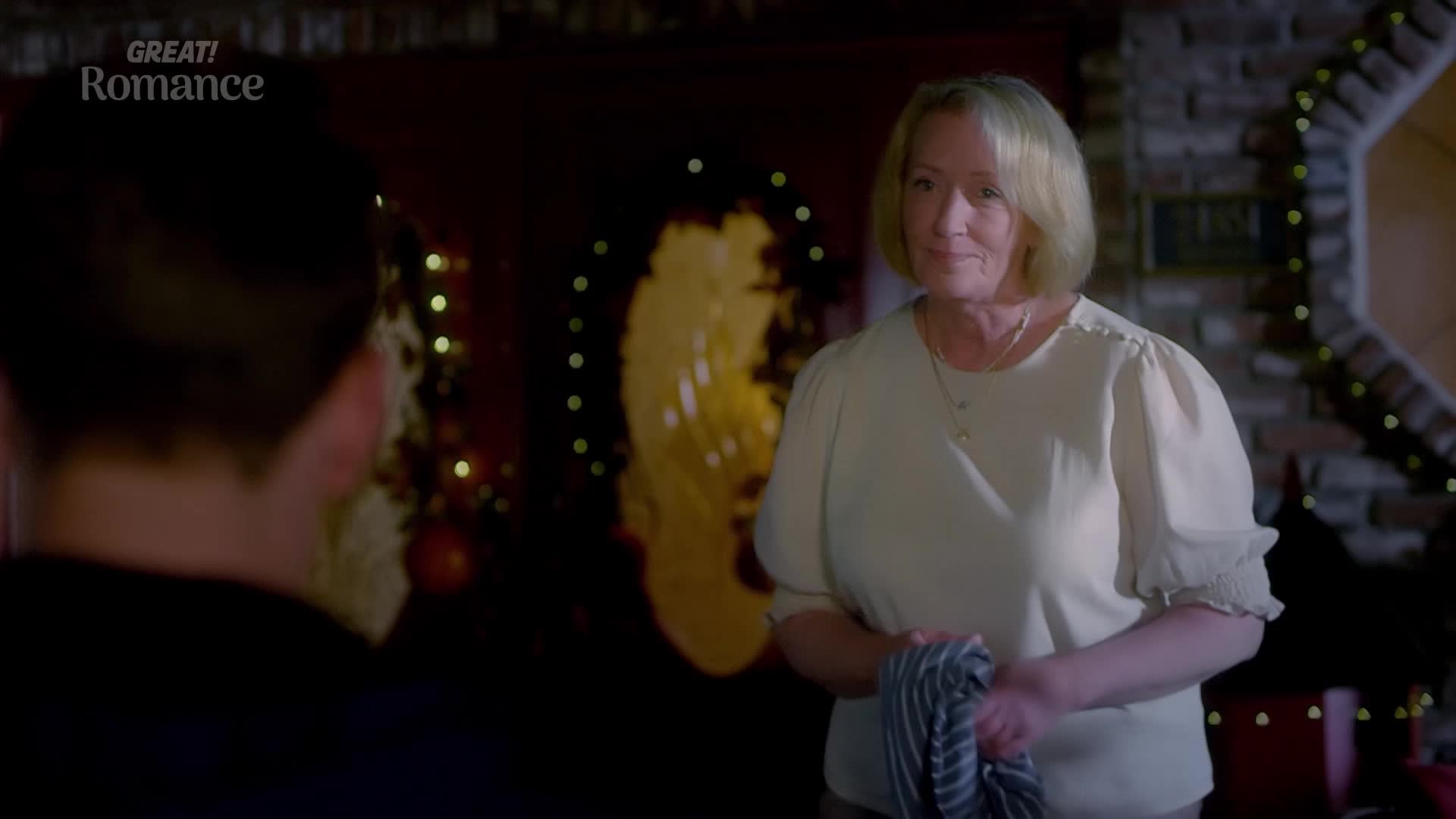 A woman with blonde hair holds a striped tea towel, smiling gently at someone off-camera. Behind her, twinkling lights and festive decorations suggest a cozy setting, perhaps a home in the UK.
