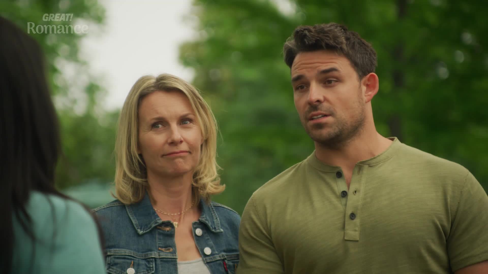 The woman in the denim jacket looks a bit skeptical, while the man in the green shirt seems to be explaining something. They're both looking at someone off-camera.
