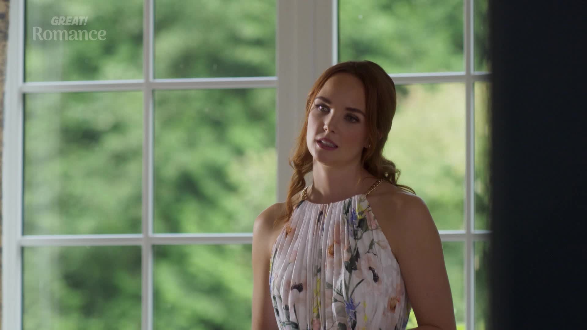 She stands in a bright room, the floral pattern of her dress contrasting with the green outside the window. Her gaze is focused, as if listening intently to someone just out of view.
