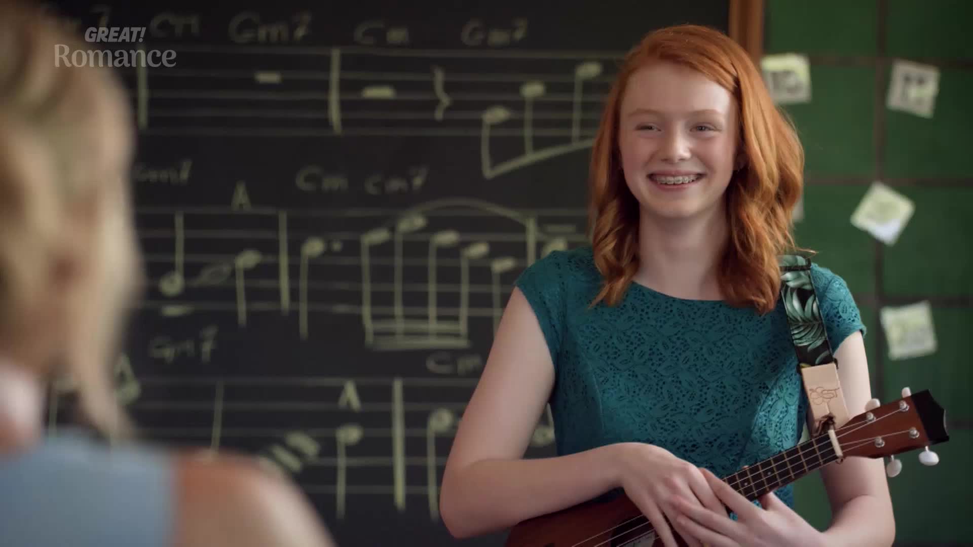 A young woman with bright red hair grins, holding a ukulele in a classroom. Behind her, a chalkboard is covered in musical notation.
