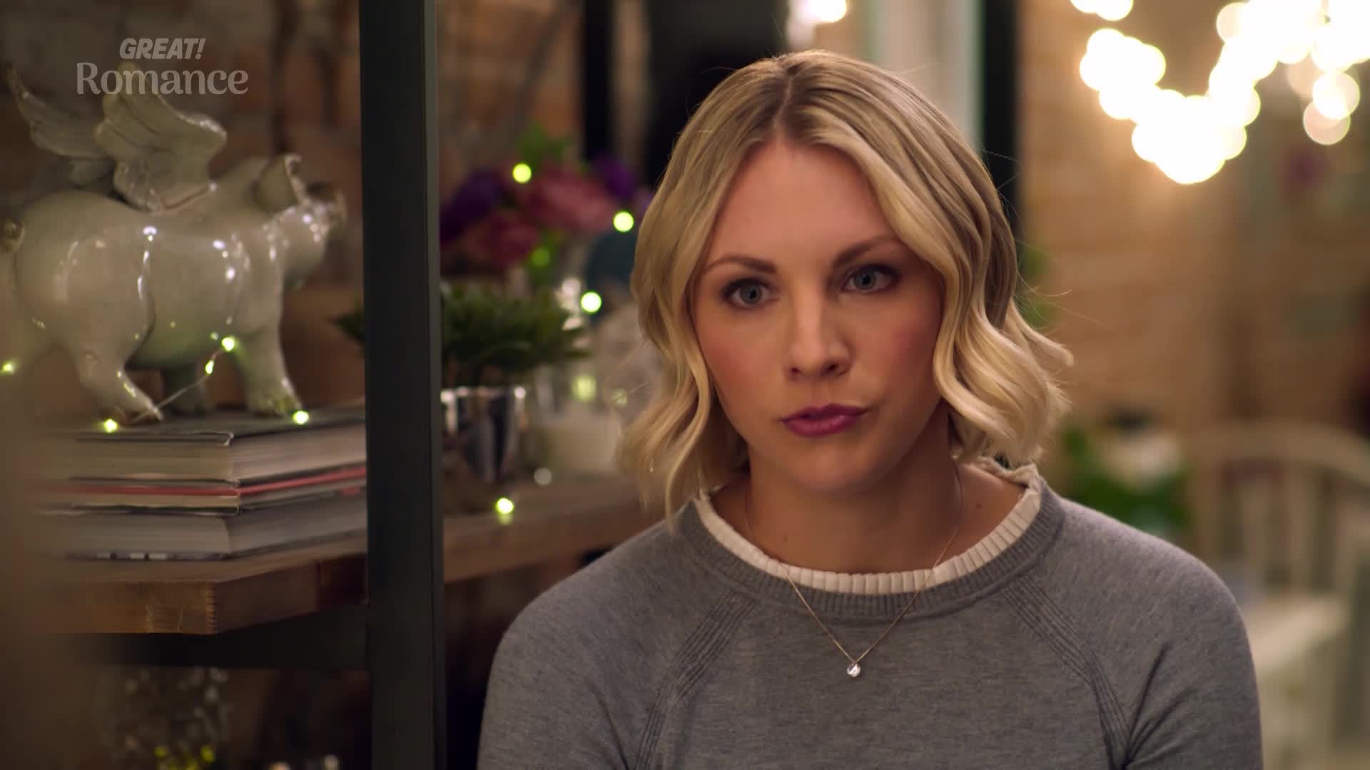 A woman with blonde waves looks directly forward, her expression thoughtful. Behind her, a shelf holds a decorative pig with wings and a string of tiny lights, hinting at a cozy, romantic setting.
