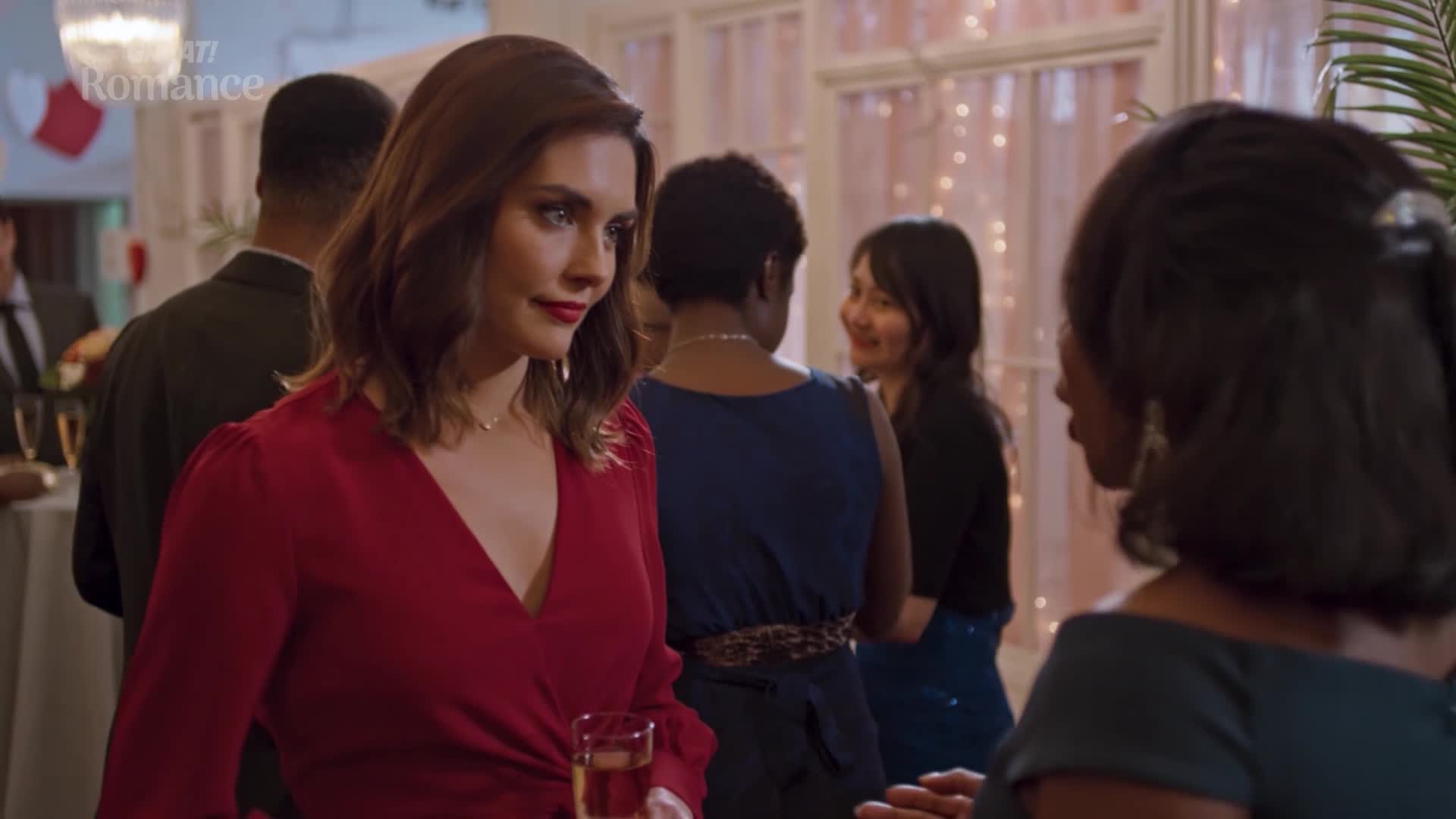 A woman in a red dress, holding a glass, is looking intently at another woman. Around them, people are chatting and mingling at what looks like a festive gathering in a well-lit room.
