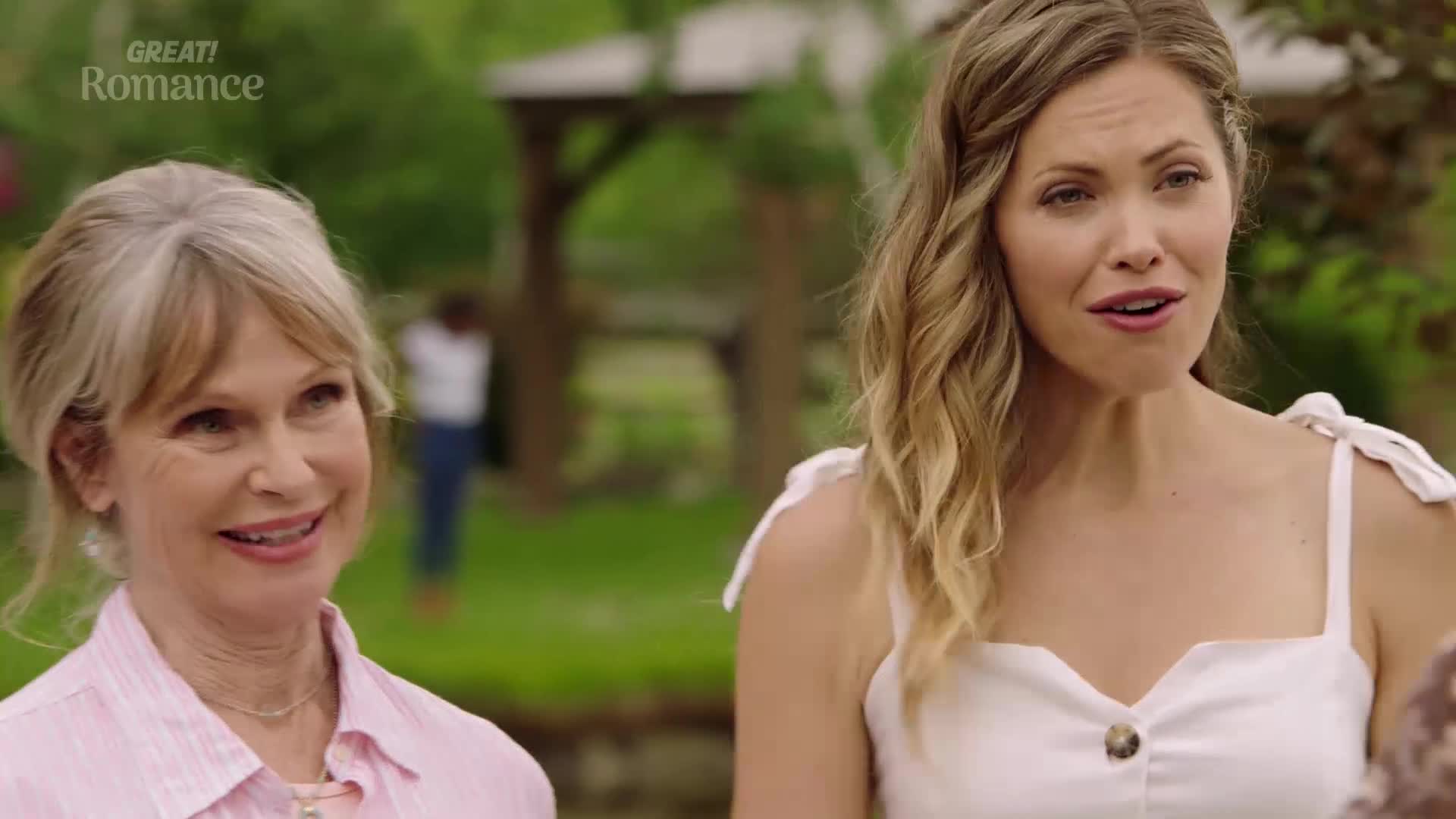 A woman in a white dress speaks animatedly, her expression open. Beside her, another woman in a pink shirt smiles, listening intently in a garden setting.
