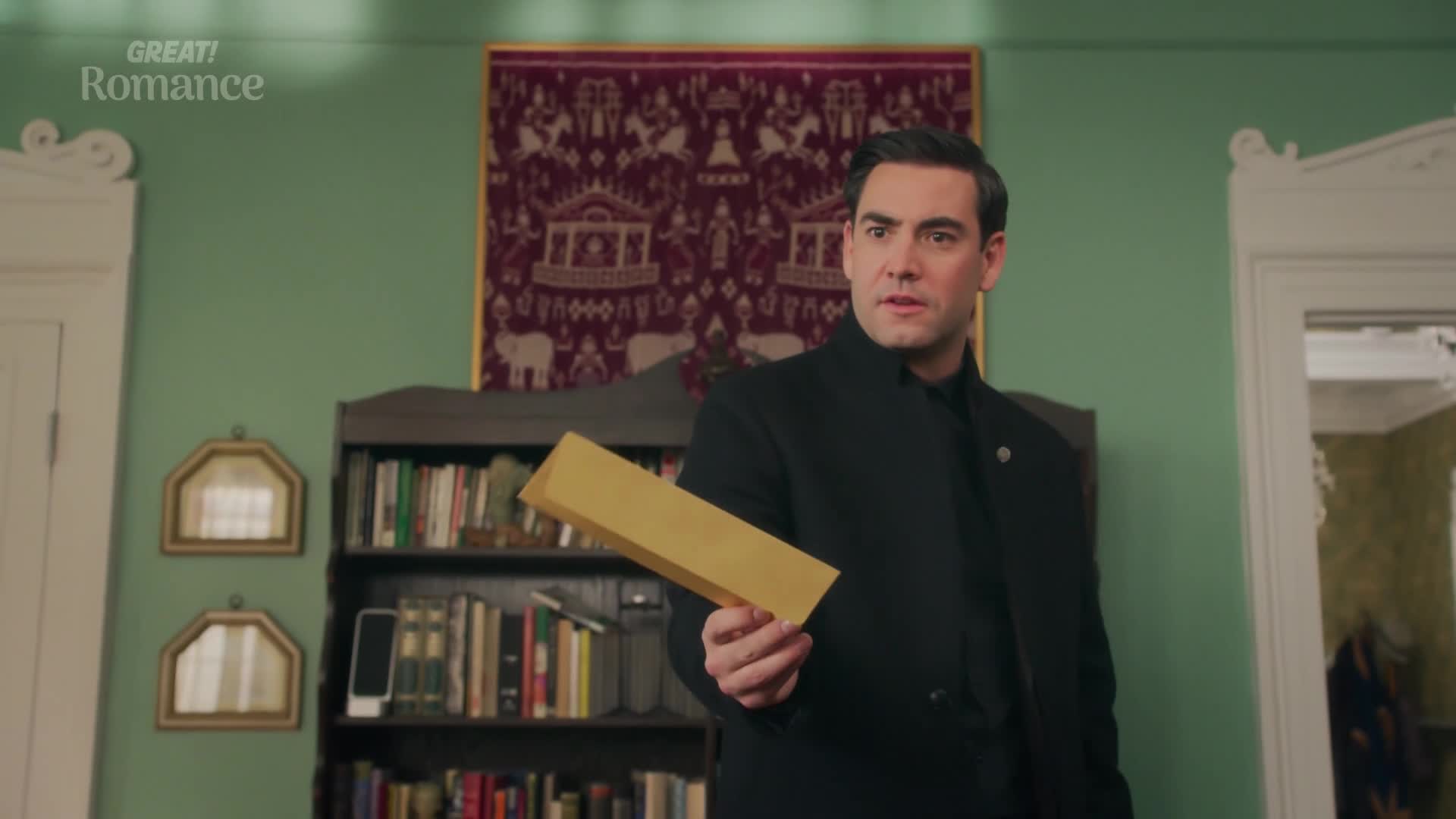 The man, dressed in black, holds a long, golden envelope, his gaze fixed on something off-camera. He stands in a room with a bookshelf and a tapestry, the setting suggesting a scene from a GREAT! romance.
