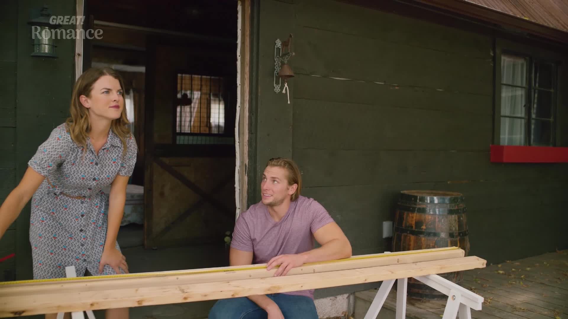 A woman in a floral dress gestures while looking up, as a man with blond hair sits, measuring a wooden plank. They're outside a barn, likely working on a project together.
