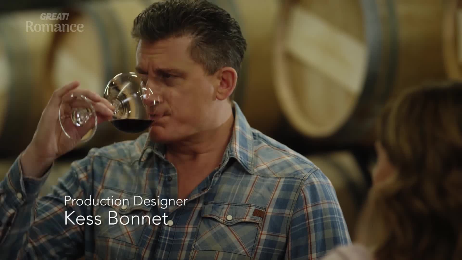 He's tilting his head back, taking a slow sip from a wine glass. The man in the plaid shirt is in a cellar, surrounded by large wooden barrels.
