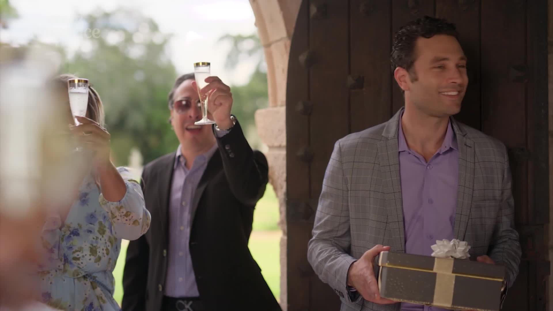 A man in a grey blazer, holding a gift, smiles as someone raises a champagne flute in a toast. Behind him, another man in a suit and sunglasses also holds a glass aloft, celebrating.
