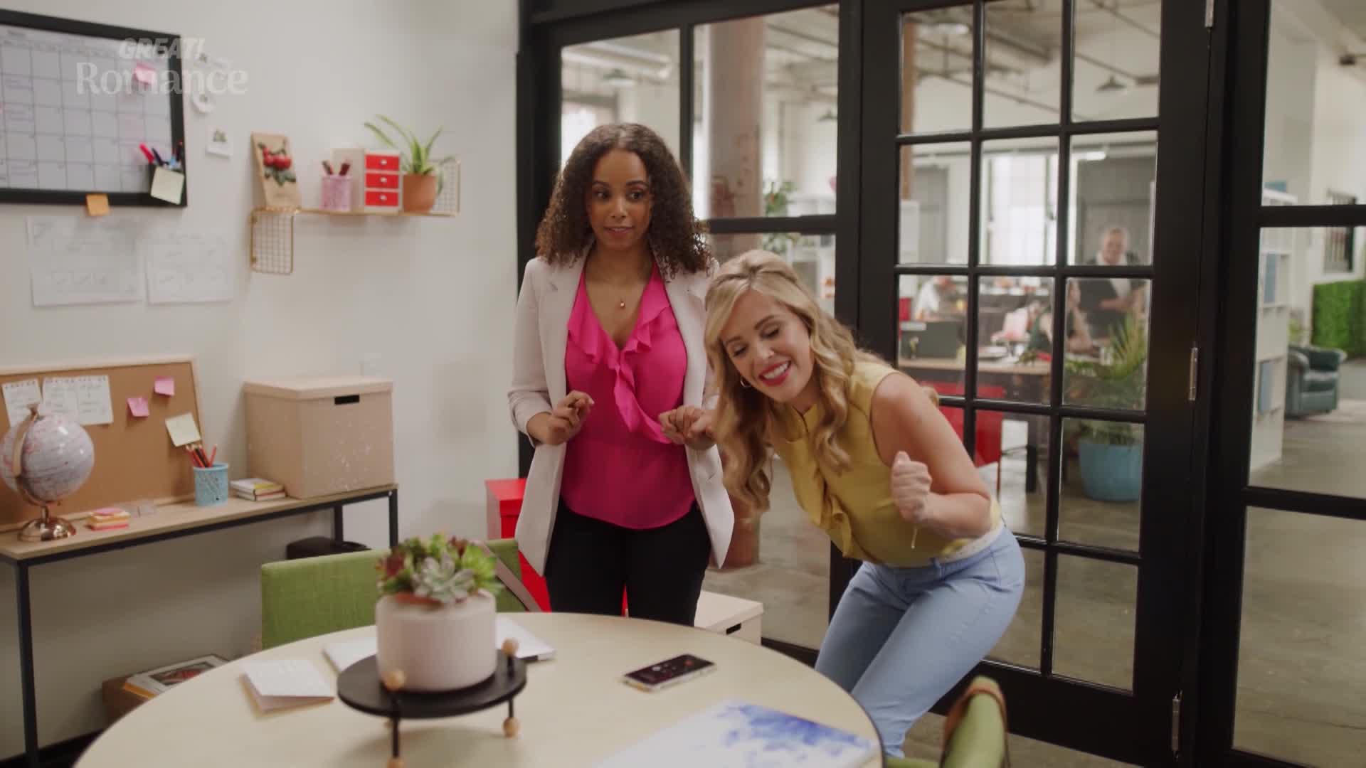 Two women are celebrating in an office, one with her hands clasped in front of her chest, the other with her fists raised in excitement. The "GREAT! Romance" logo is visible in the upper left corner.