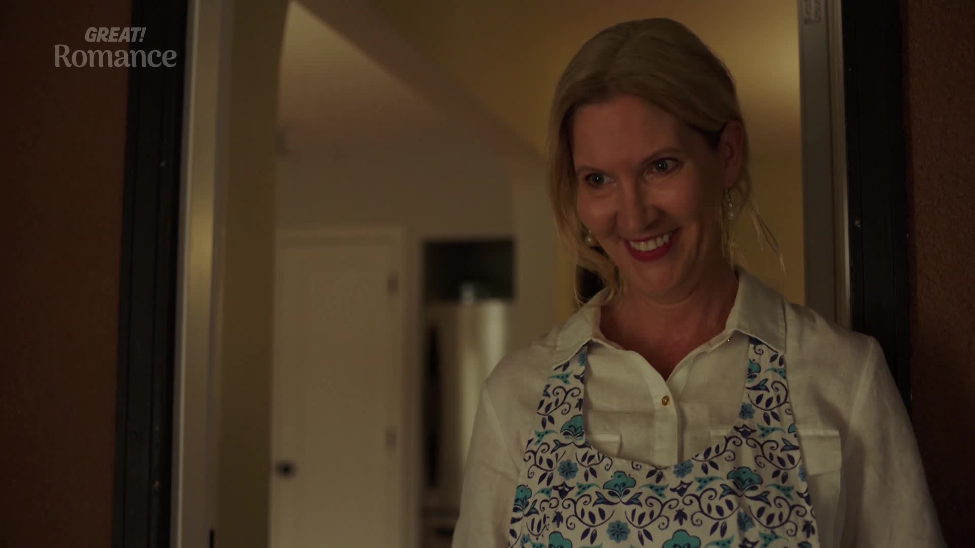 A woman with blonde hair smiles warmly, her eyes crinkling at the corners. She wears a white shirt and a blue floral apron, standing just inside a doorway. A woman with blonde hair smiles warmly, her eyes crinkling at the corners. She wears a white shirt and a blue floral apron, standing just inside a doorway.