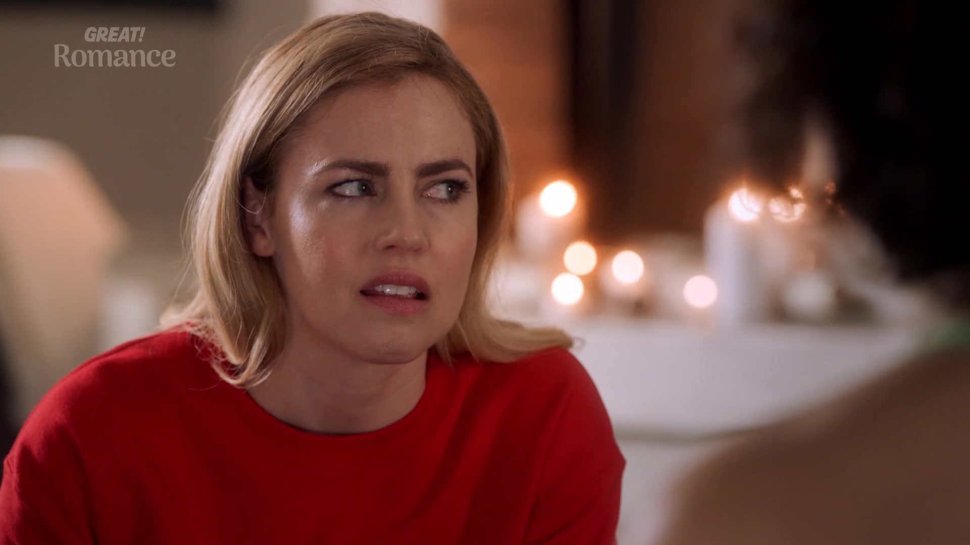 A woman with blonde hair, wearing a bright red jumper, looks off to the side with a worried expression. Behind her, soft lights glow, hinting at a cozy evening indoors. A woman with blonde hair, wearing a bright red jumper, looks off to the side with a worried expression. Behind her, soft lights glow, hinting at a cozy evening indoors.