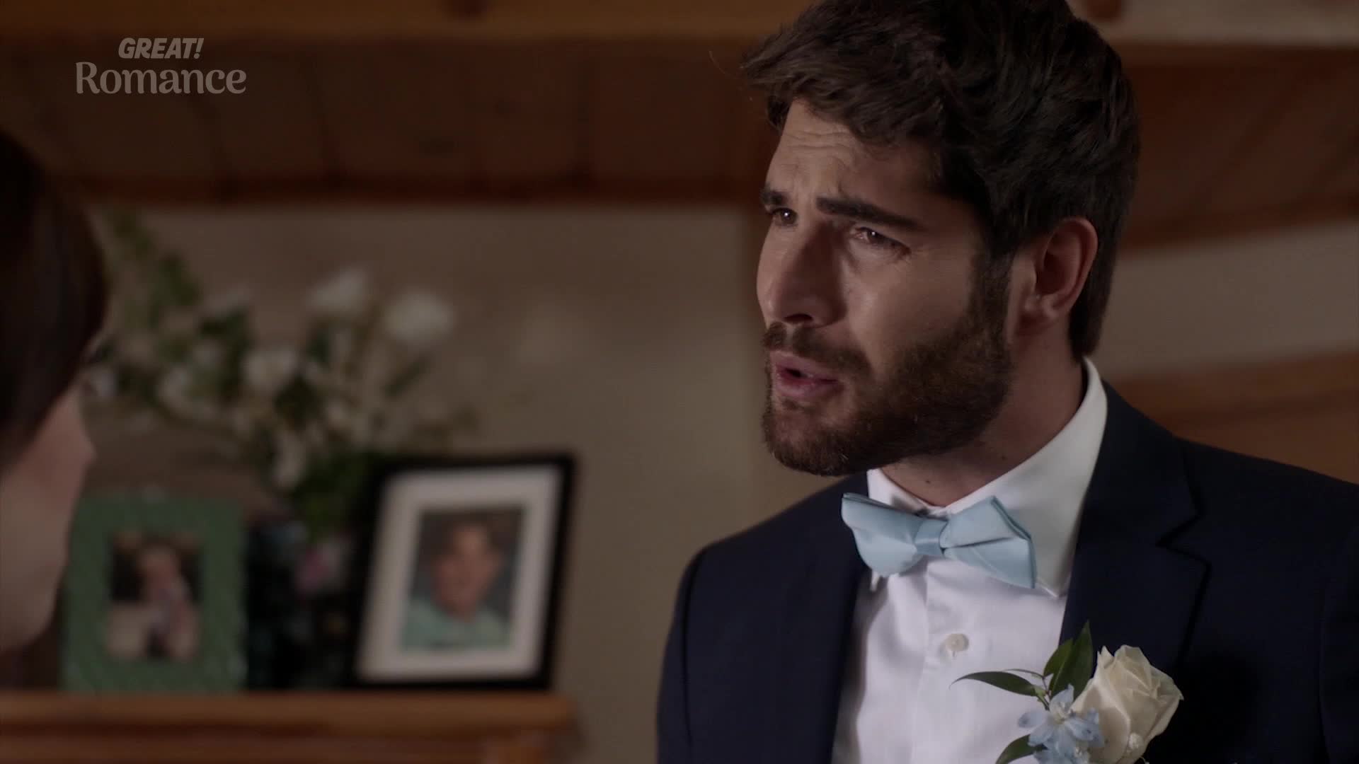 A man in a navy suit and a light blue bowtie speaks with a furrowed brow. Framed photos sit on a shelf behind him, hinting at a life in the UK. A man in a navy suit and a light blue bowtie speaks with a furrowed brow. Framed photos sit on a shelf behind him, hinting at a life in the UK.