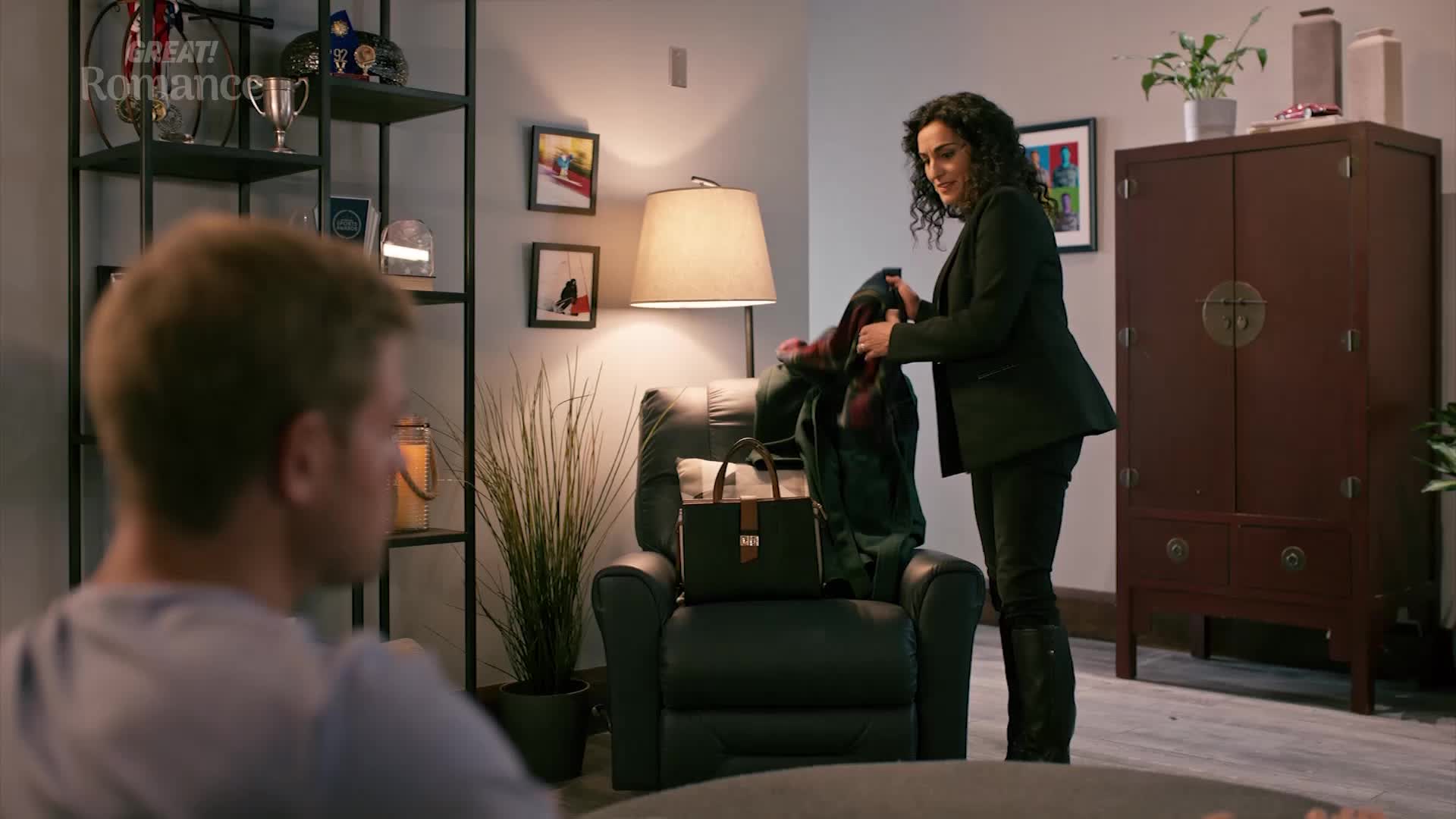 A woman in a dark suit is packing a tartan scarf into a green jacket. She stands by a recliner, a leather bag resting on its seat. A woman in a dark suit is packing a tartan scarf into a green jacket. She stands by a recliner, a leather bag resting on its seat.