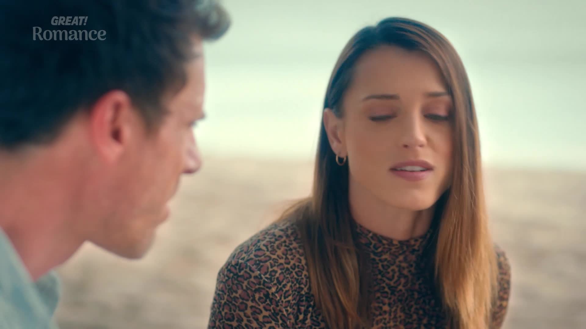 A man in a light blue shirt leans in, his gaze fixed on the woman beside him. She, wearing a leopard-print top, closes her eyes as if lost in thought.