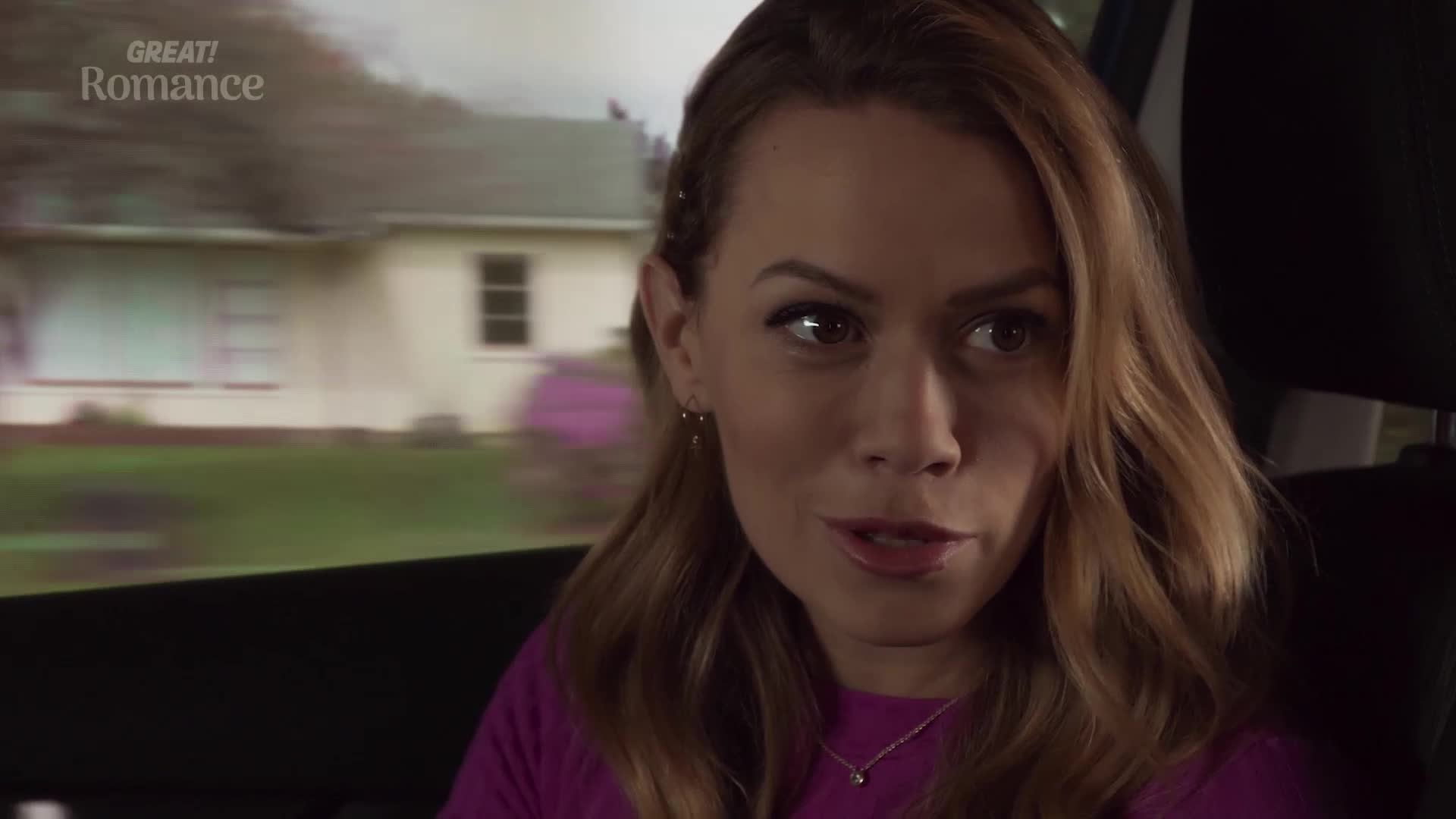 A woman with blonde hair is talking, her eyes bright. Outside the car window, houses and trees streak by.