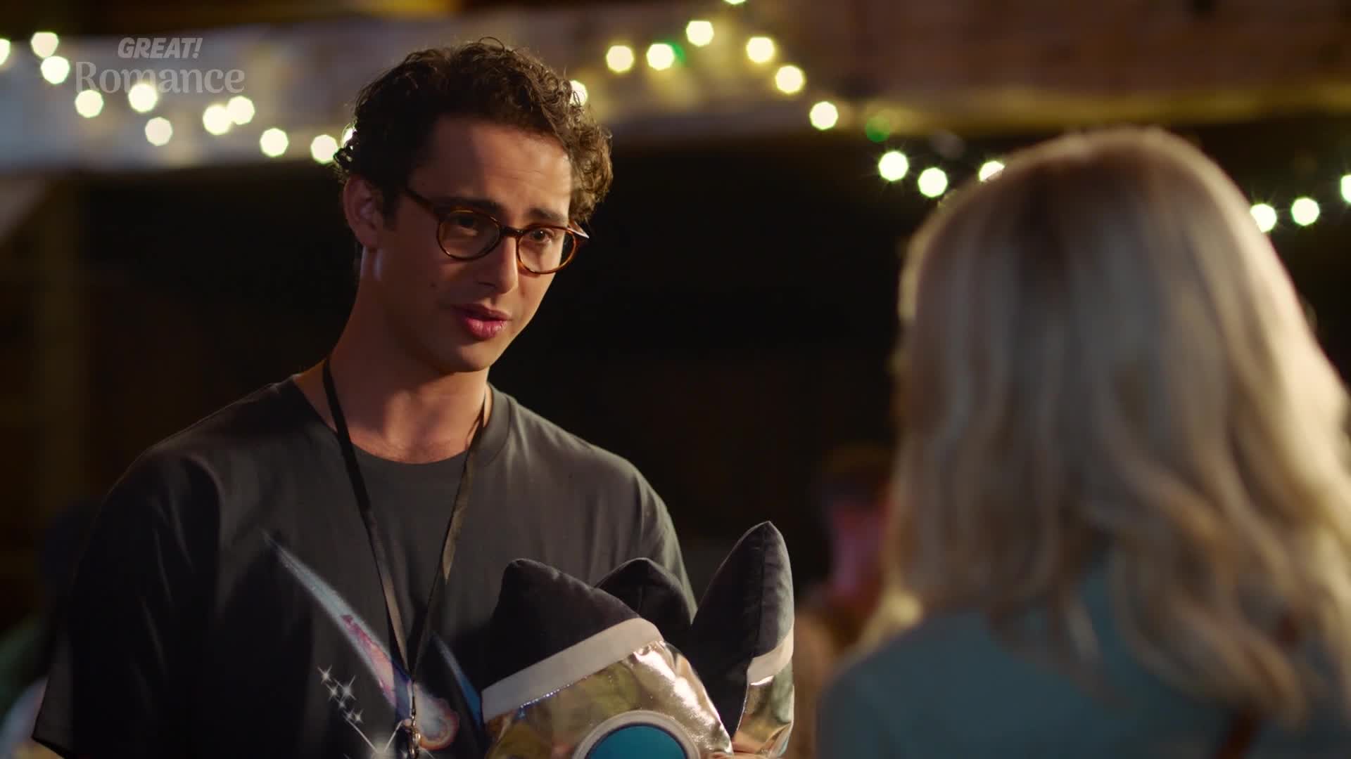 A young man with glasses gestures with a plush toy, its metallic gold and black fabric catching the light. He's speaking to a woman with blonde hair, her back to me, under a string of fairy lights.
