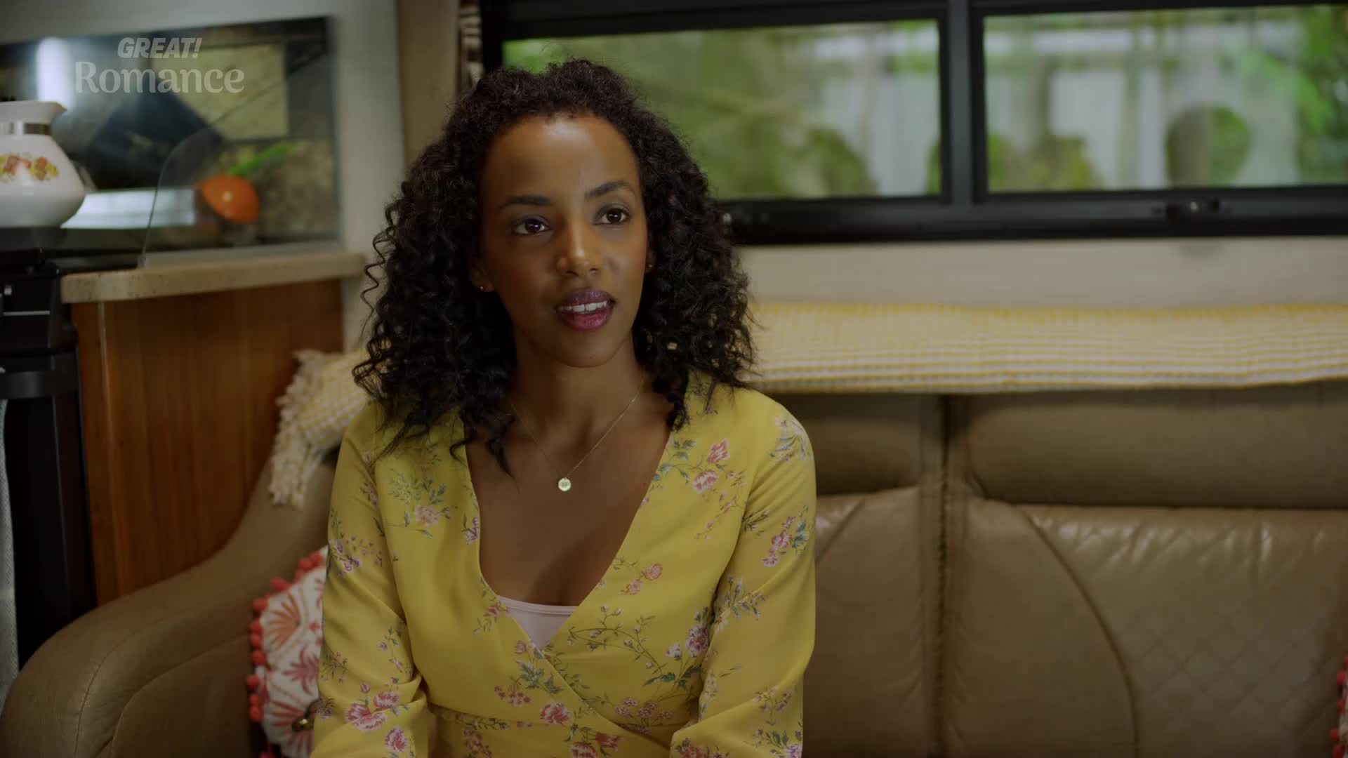 A woman with curly dark hair, wearing a yellow floral dress, sits on a brown sofa inside what appears to be a caravan. Sunlight filters through a window behind her, illuminating the cozy interior.