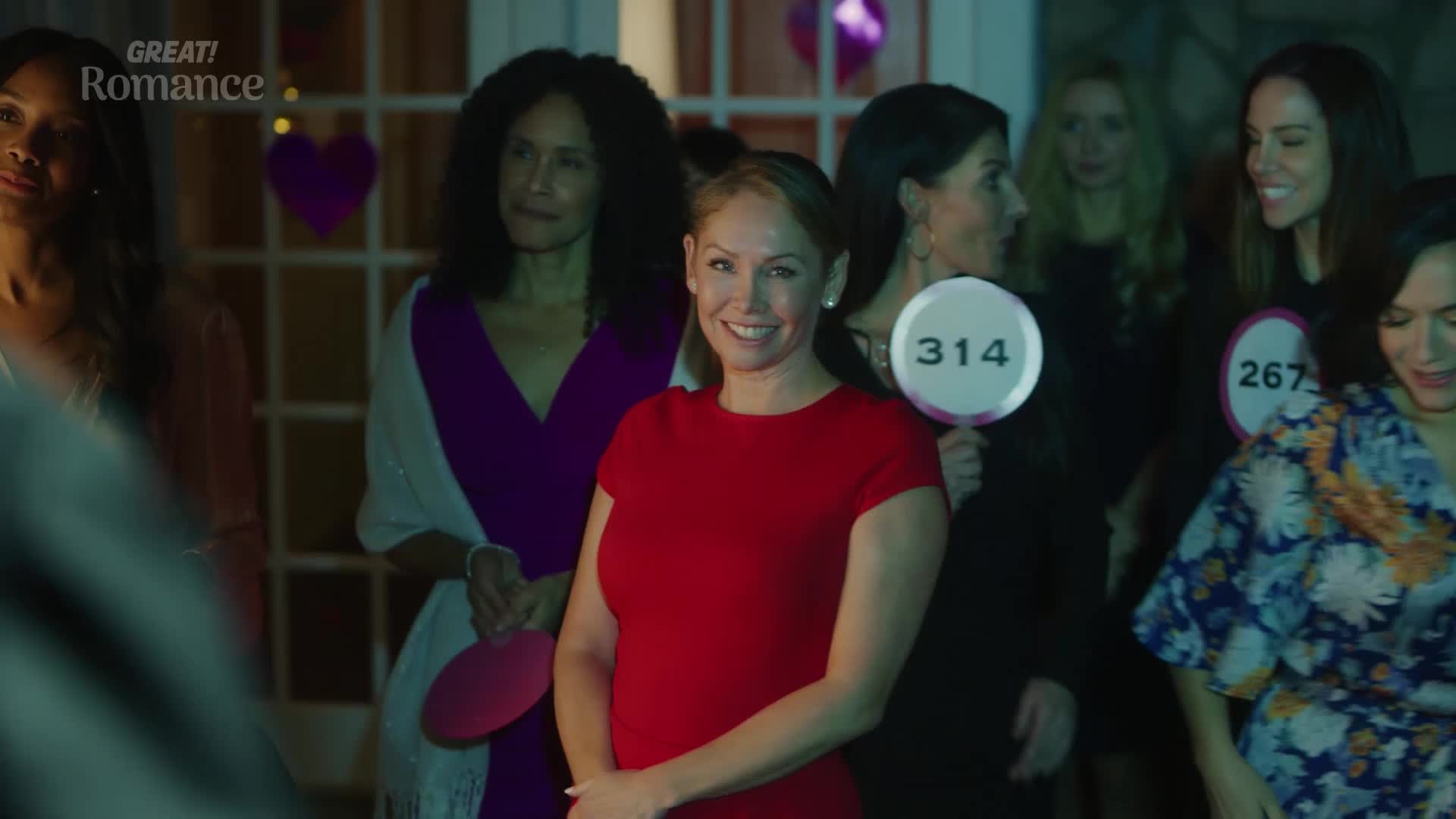 A woman in a bright red dress smiles, holding a paddle with "314" on it. Behind her, other women are gathered, some also holding paddles, at what looks like a lively charity auction.