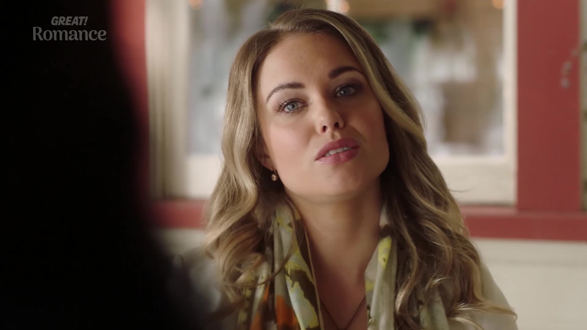 A woman with blonde, wavy hair speaks, her eyes meeting mine. She wears a patterned scarf, a delicate pearl earring, and a necklace.